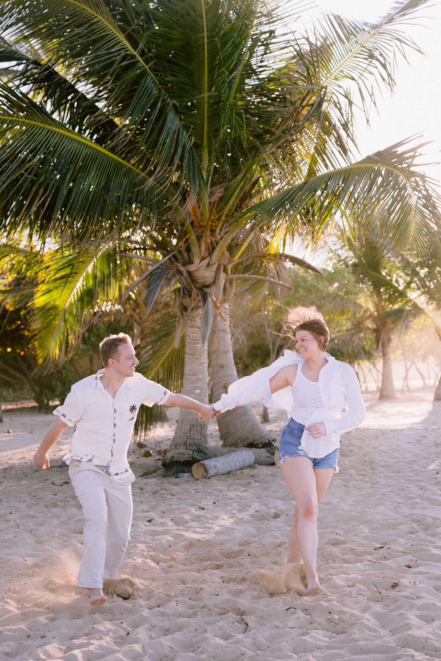 Happy couple holding hands and dancing on a tropical sandy beach surrounded by tall palm trees in golden sunlight.