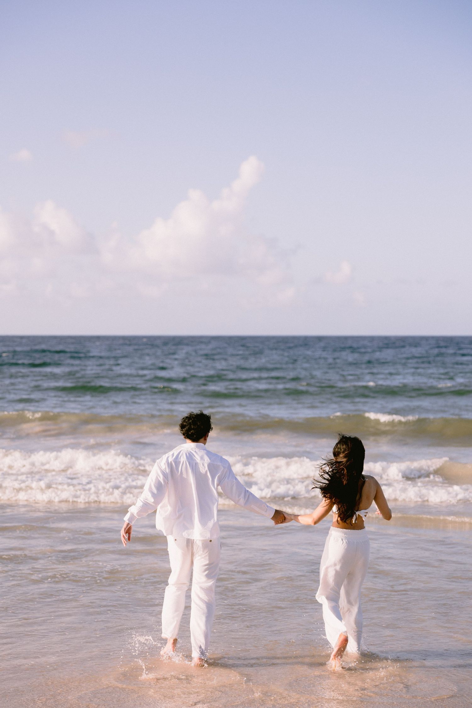 Couple in white outfits holding hands walking into ocean waves on a sunny beach.
