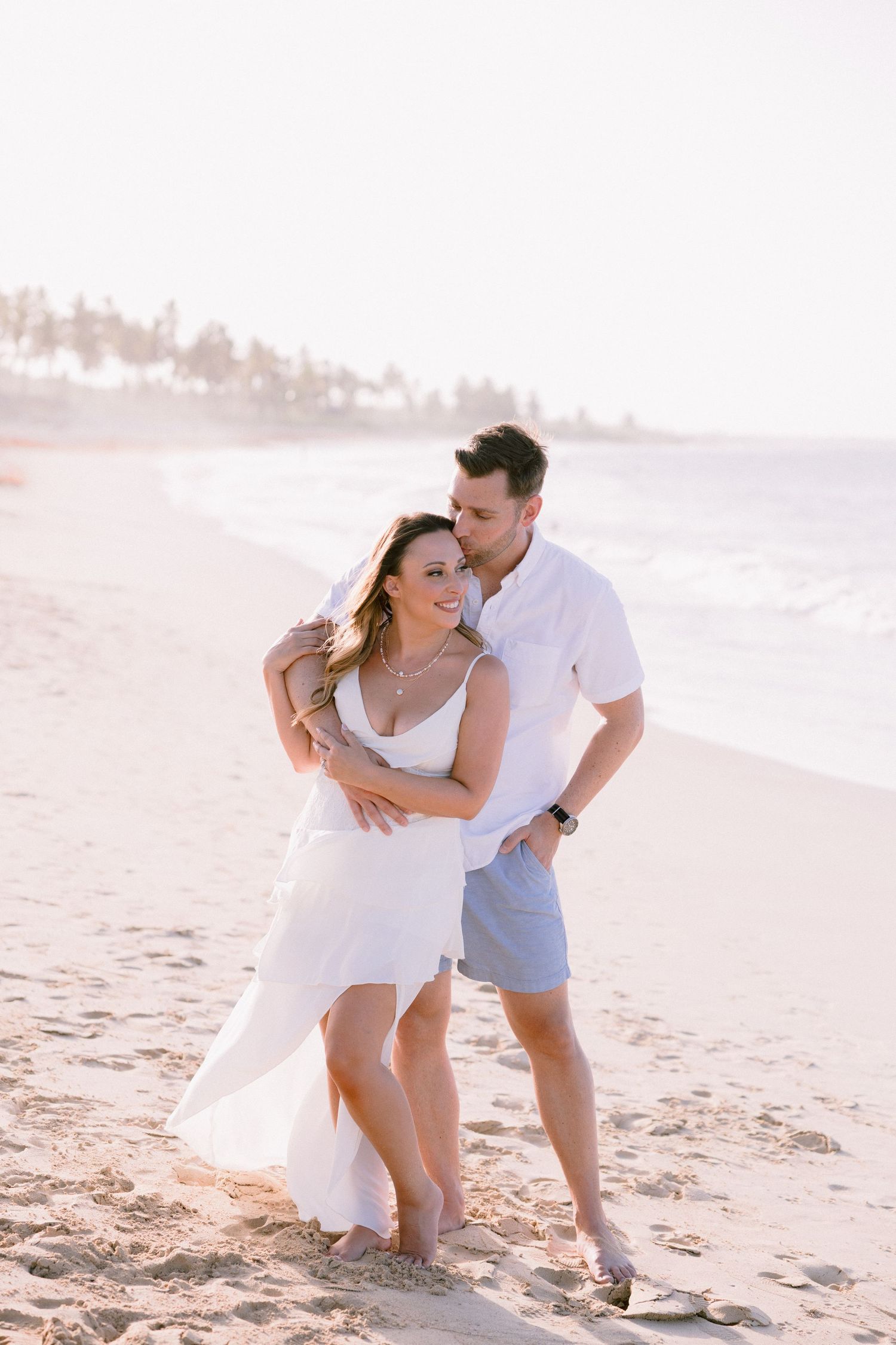 Couple in white outfits embracing on a sunny beach at golden hour with ocean waves in the background.
