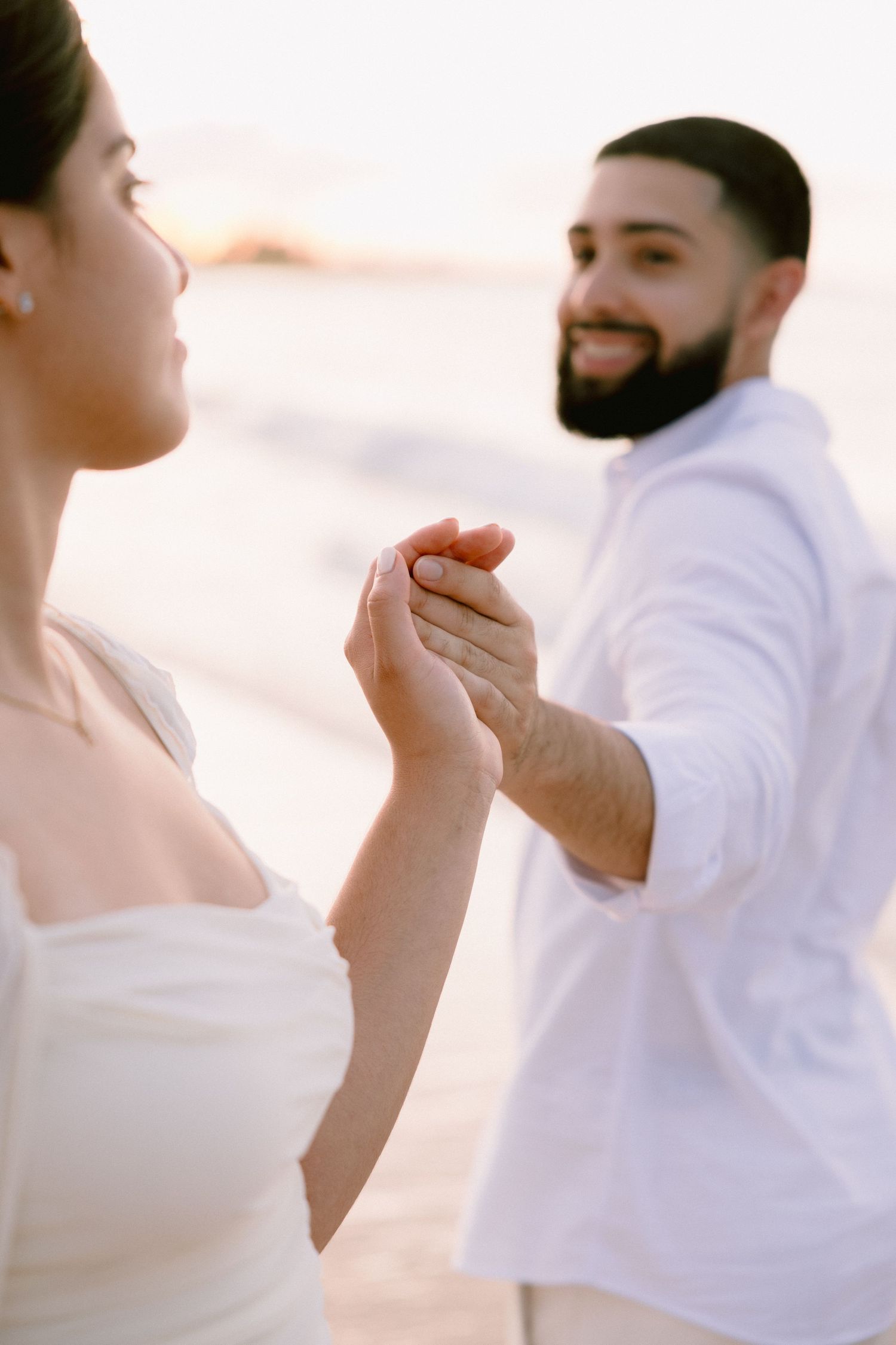 A smiling bearded man in white holds hands with a woman in a white dress on a sunlit beach.