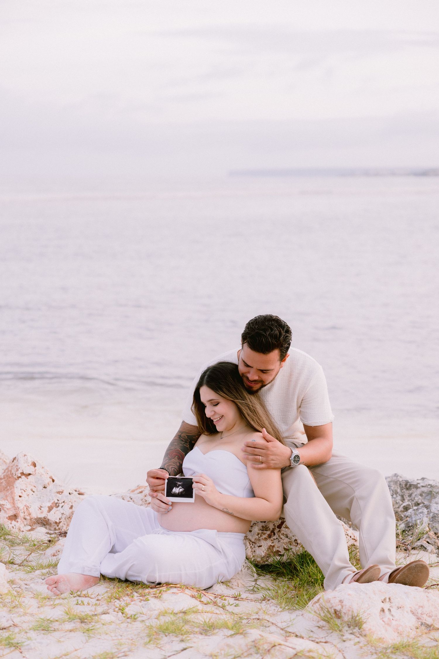 Expecting couple sitting on beach rocks, holding ultrasound photo, with calm ocean backdrop.