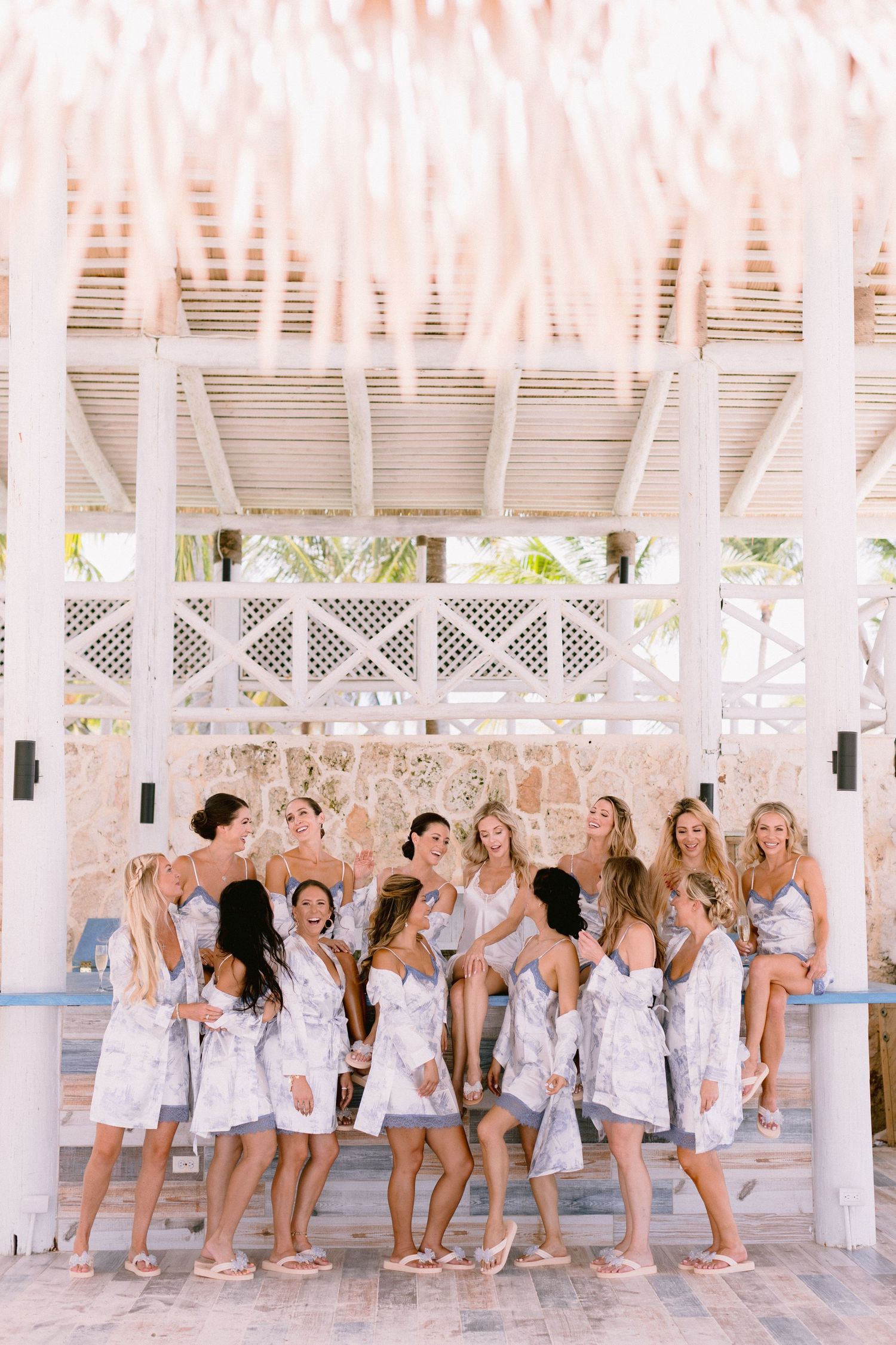Bridesmaids in matching floral robes laugh together under a white gazebo at a tropical beach wedding venue.