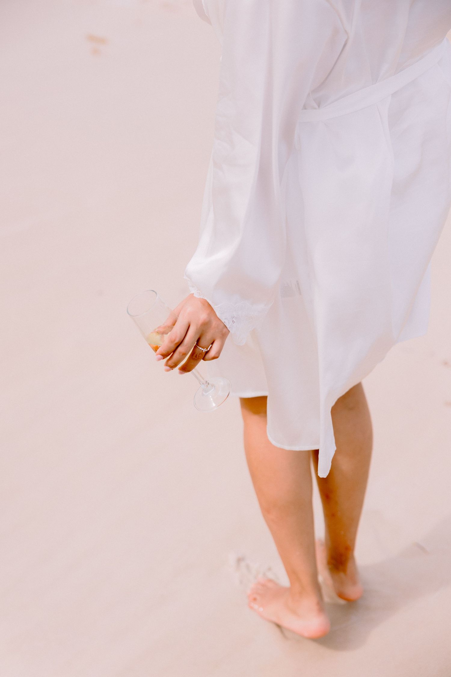 Barefoot woman in white shirt dress walking on sandy beach holding a clear glass bottle.
