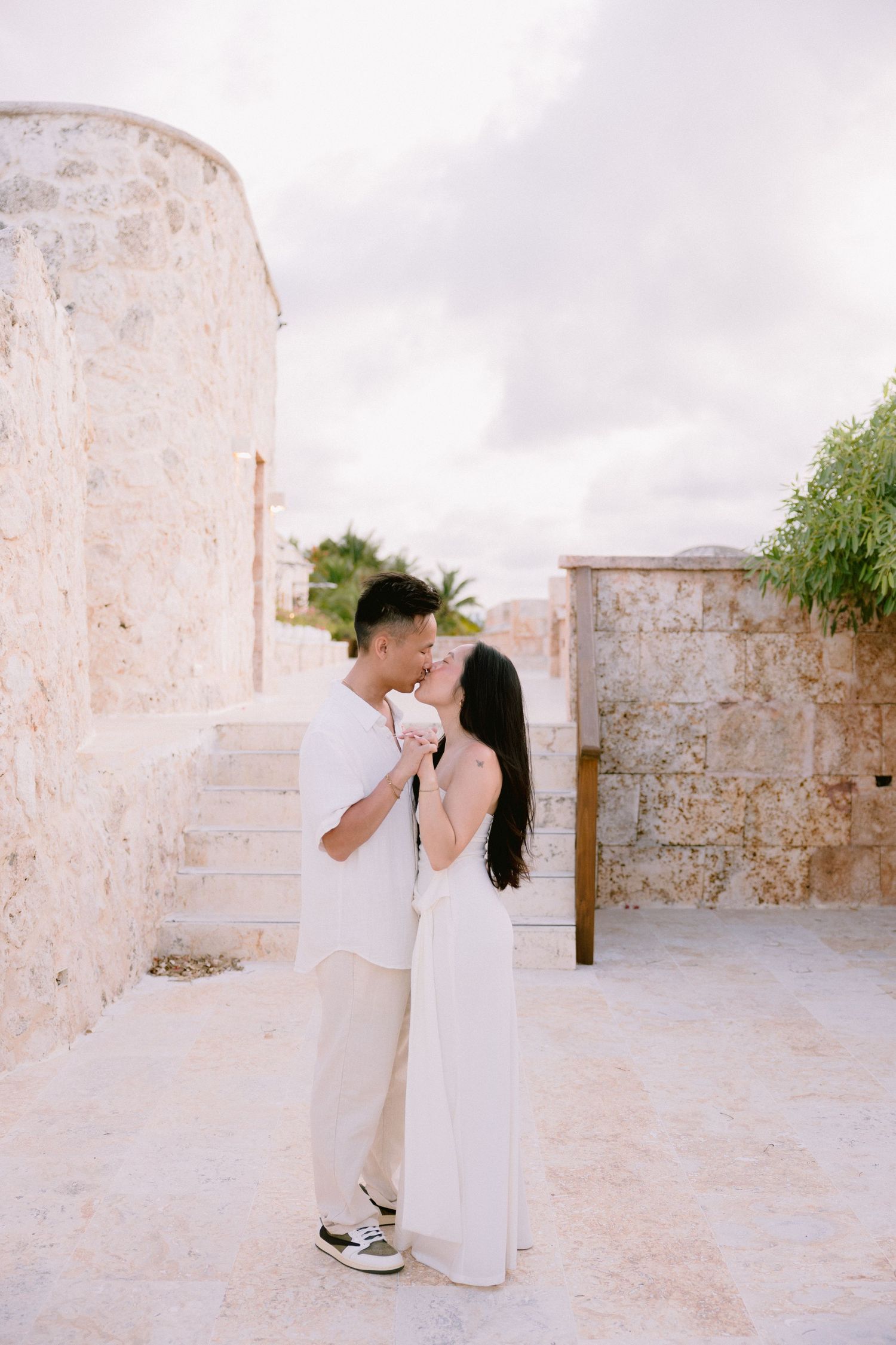 Couple sharing a romantic kiss in white outfits against a stone wall backdrop outdoors.