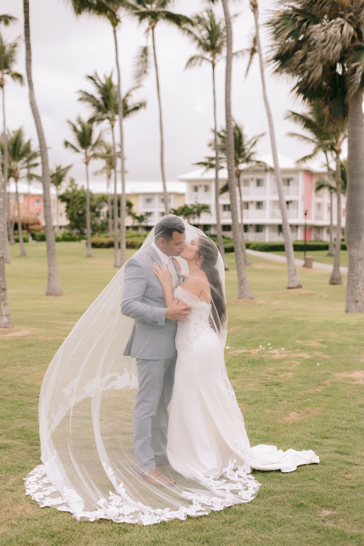 Bride and groom share a romantic kiss outdoors, with a flowing lace veil and palm trees framing their wedding portrait.