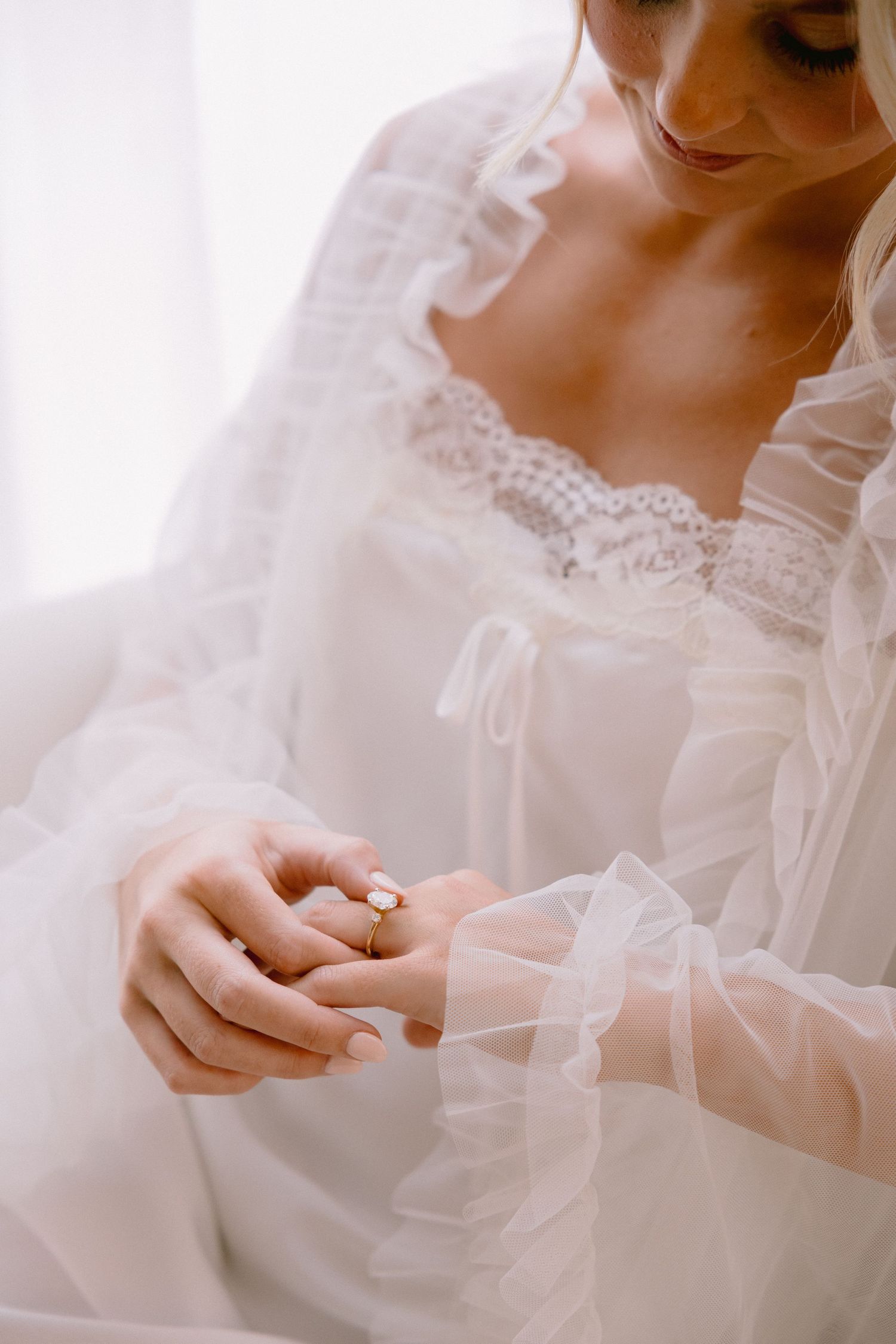 Delicate hands in white lace sleeves hold a small ring against ethereal wedding dress fabric.