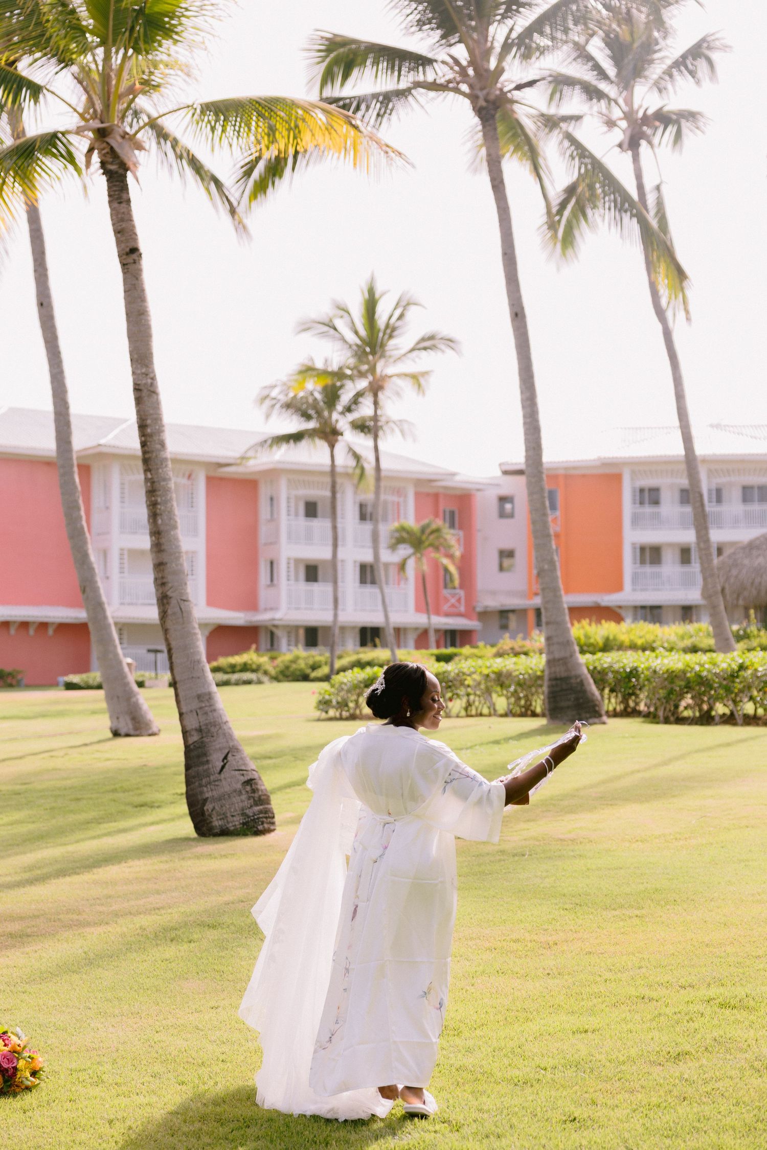 A bride in a flowing white wedding dress spins gracefully on a lush lawn with palm trees and coral buildings in the background.