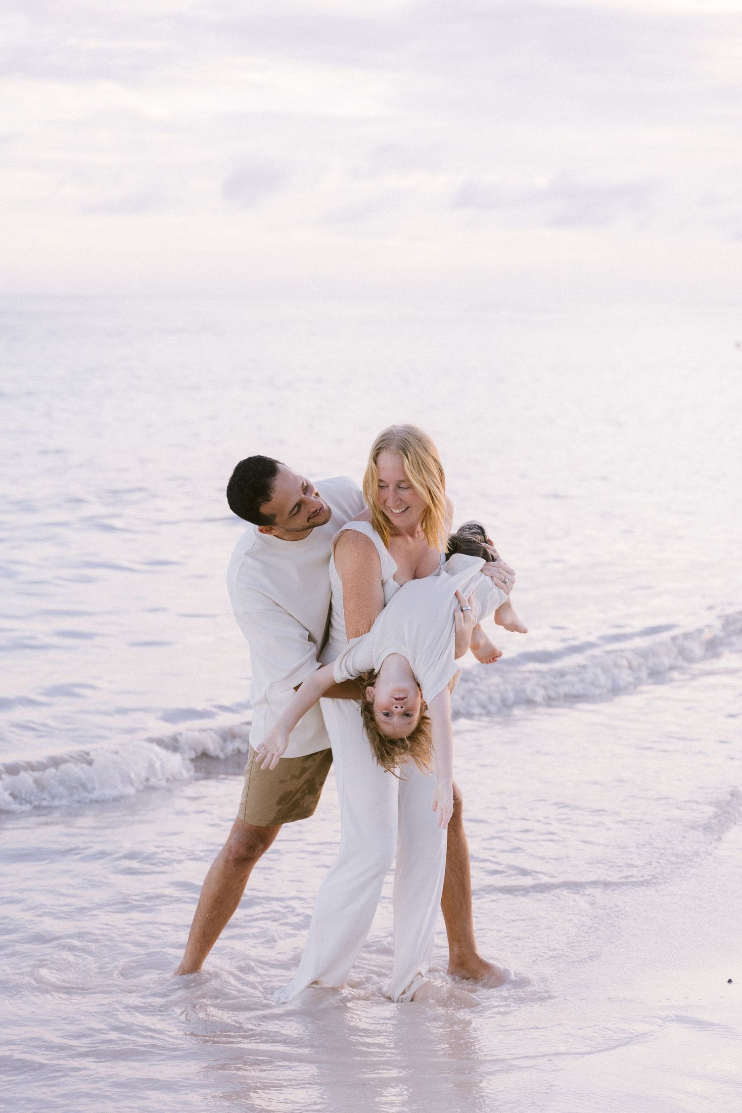 A joyful family plays on a beach at sunset, with parents swinging their laughing child upside down by the water.
