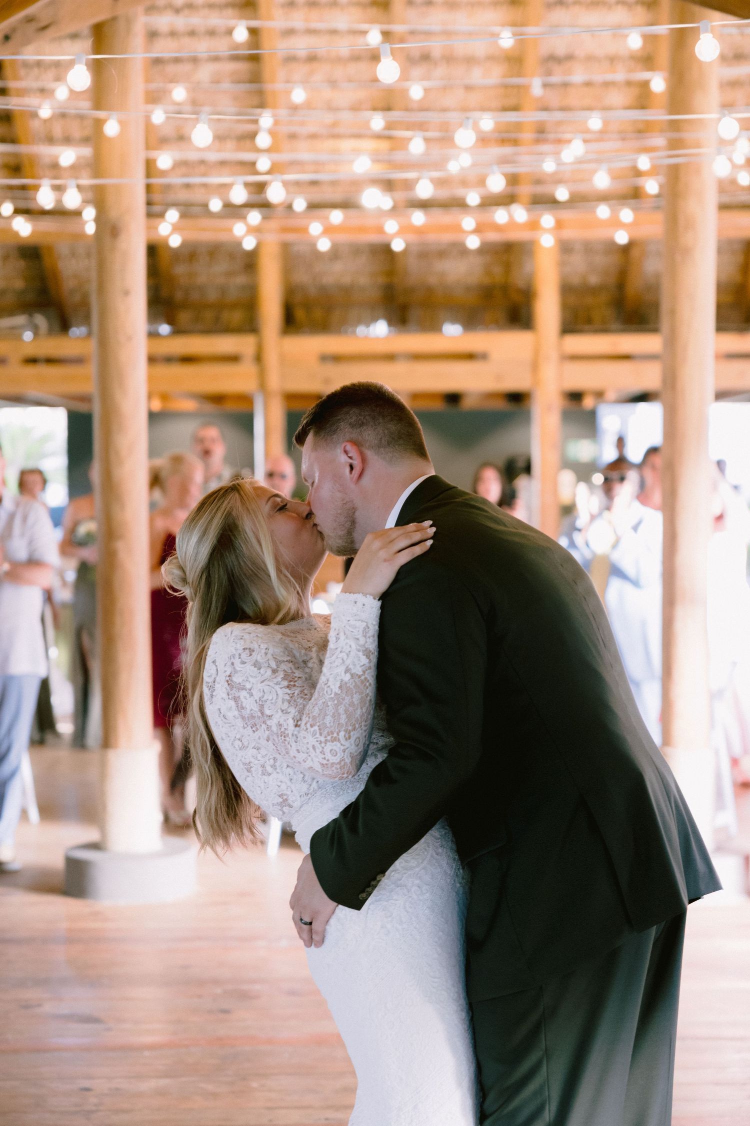 A bride and groom share their first dance kiss under warm string lights in a rustic wooden venue.