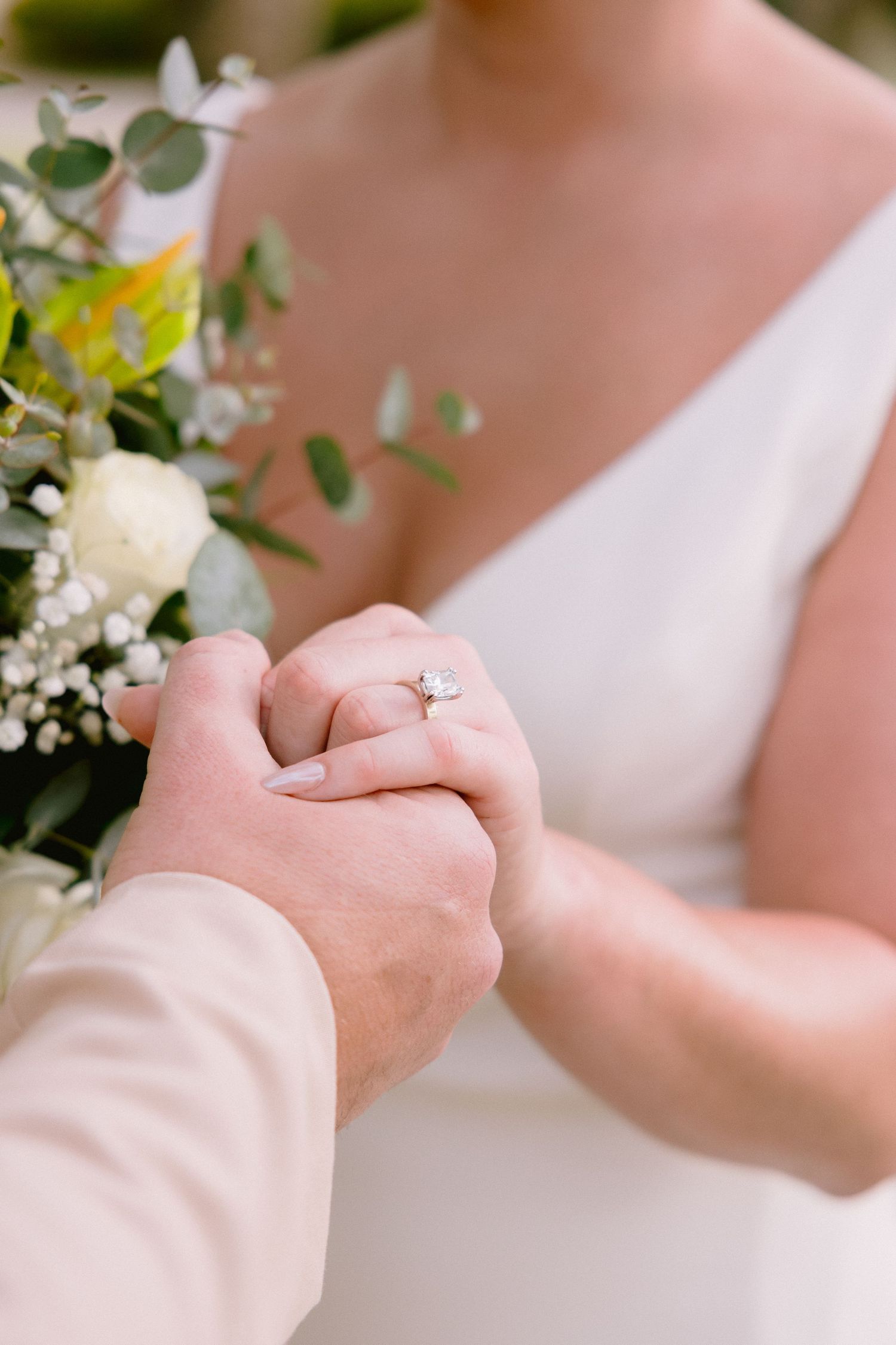 Bride and groom holding hands, showcasing a diamond engagement ring, with a white floral bouquet at a wedding ceremony.