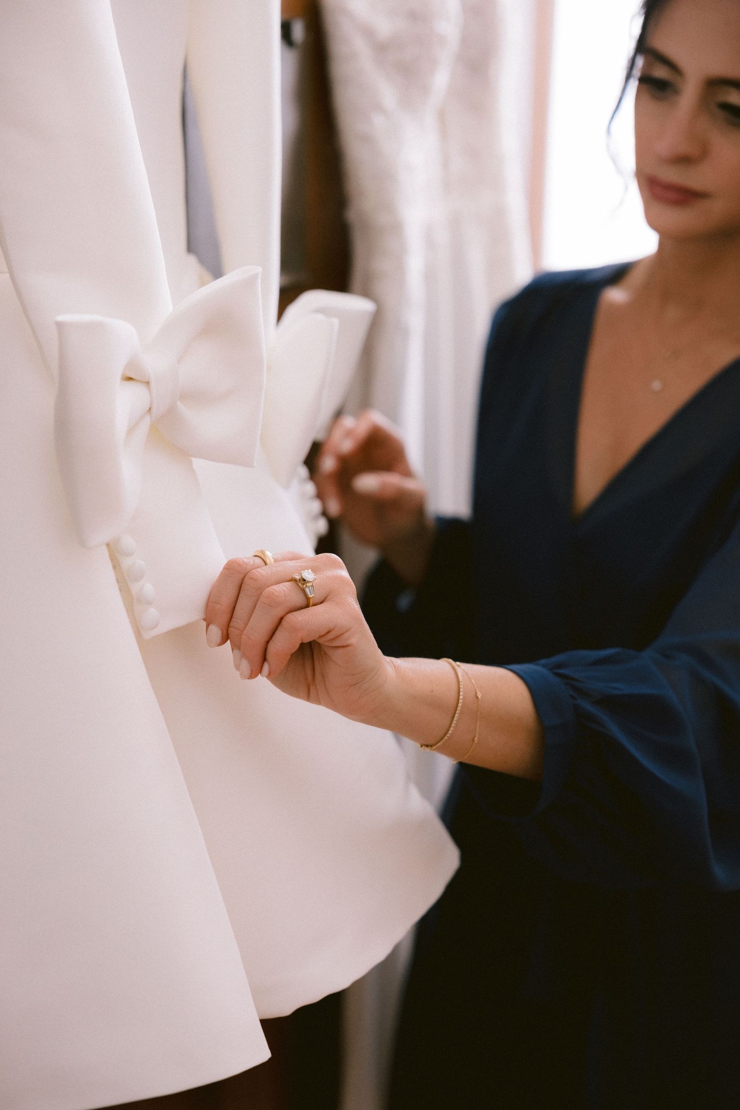 Woman in blue dress adjusting bow detail on white wedding gown hanging on rack in bridal boutique.