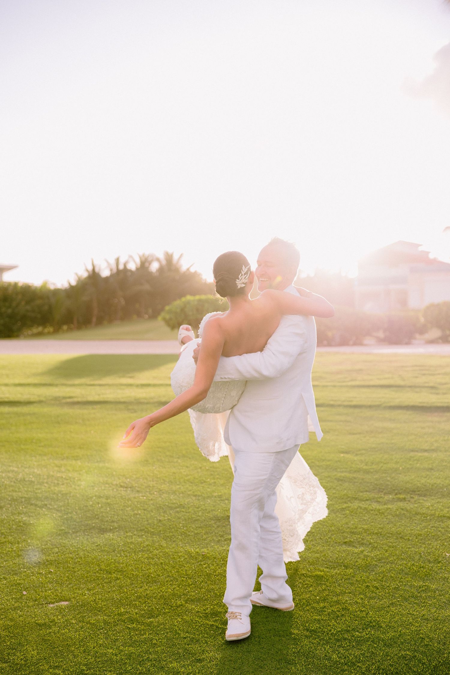 A couple in white attire dances gracefully on a sunlit grassy field during golden hour.