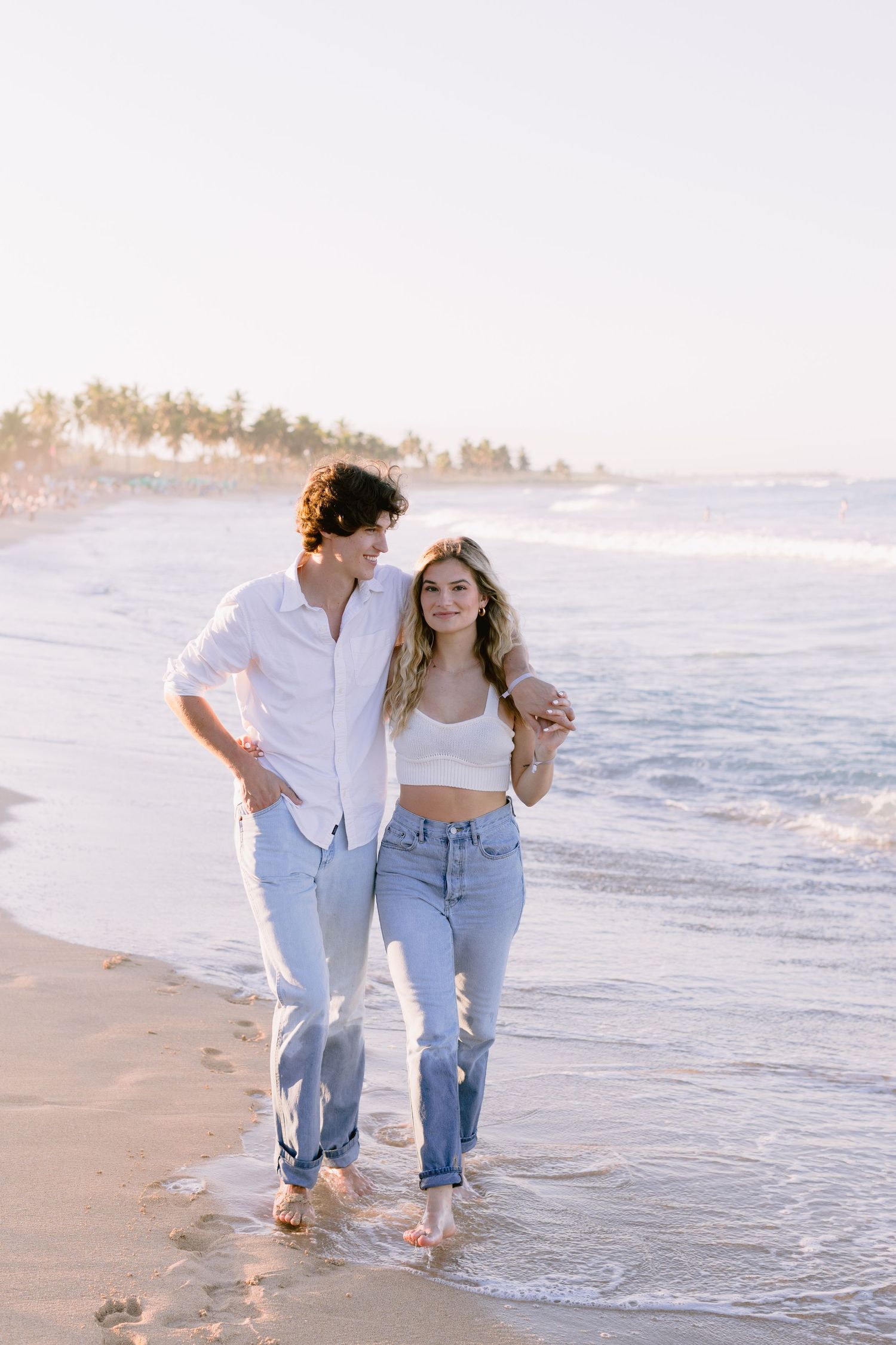 Couple in casual denim walking along the shoreline with palm trees in the background.