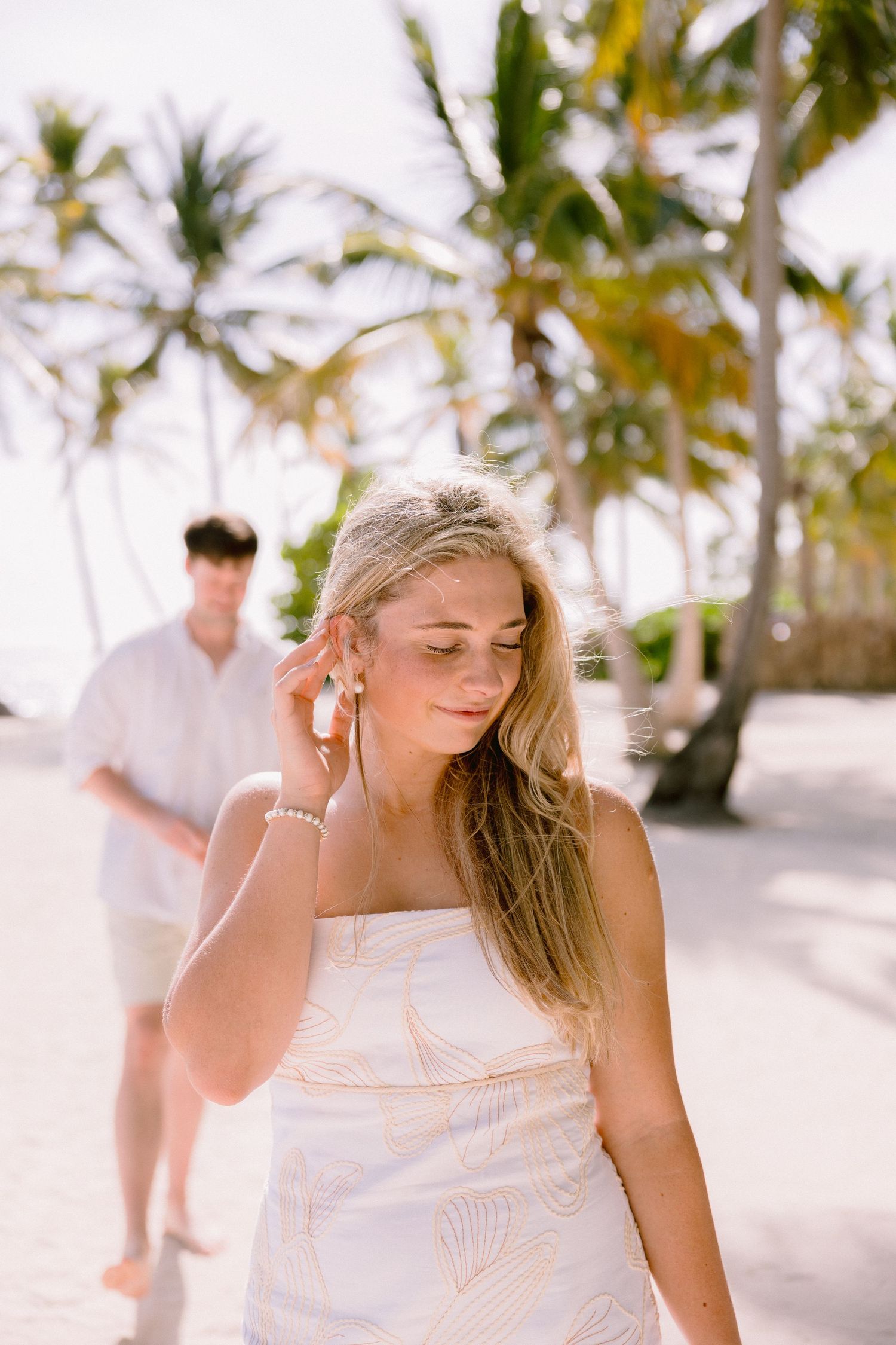 A person in a white dress walks along a tropical beach path lined with palm trees on a sunny day.