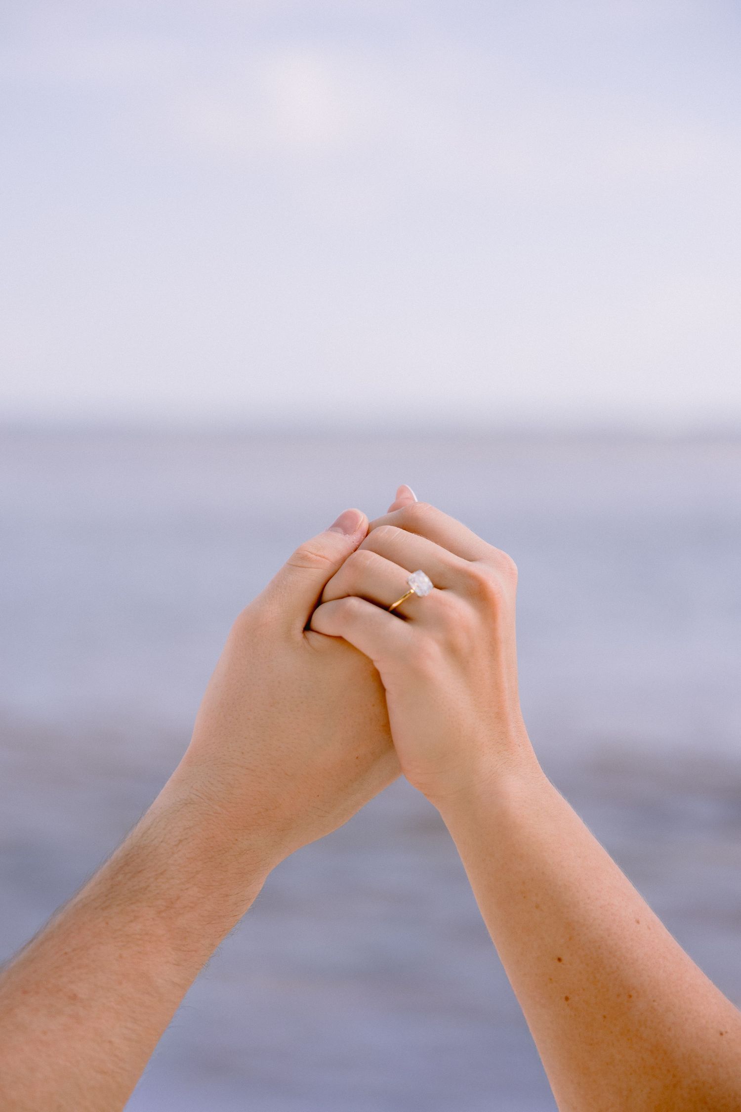 Two hands reach towards each other against a blurred ocean background, with one hand wearing an engagement ring.