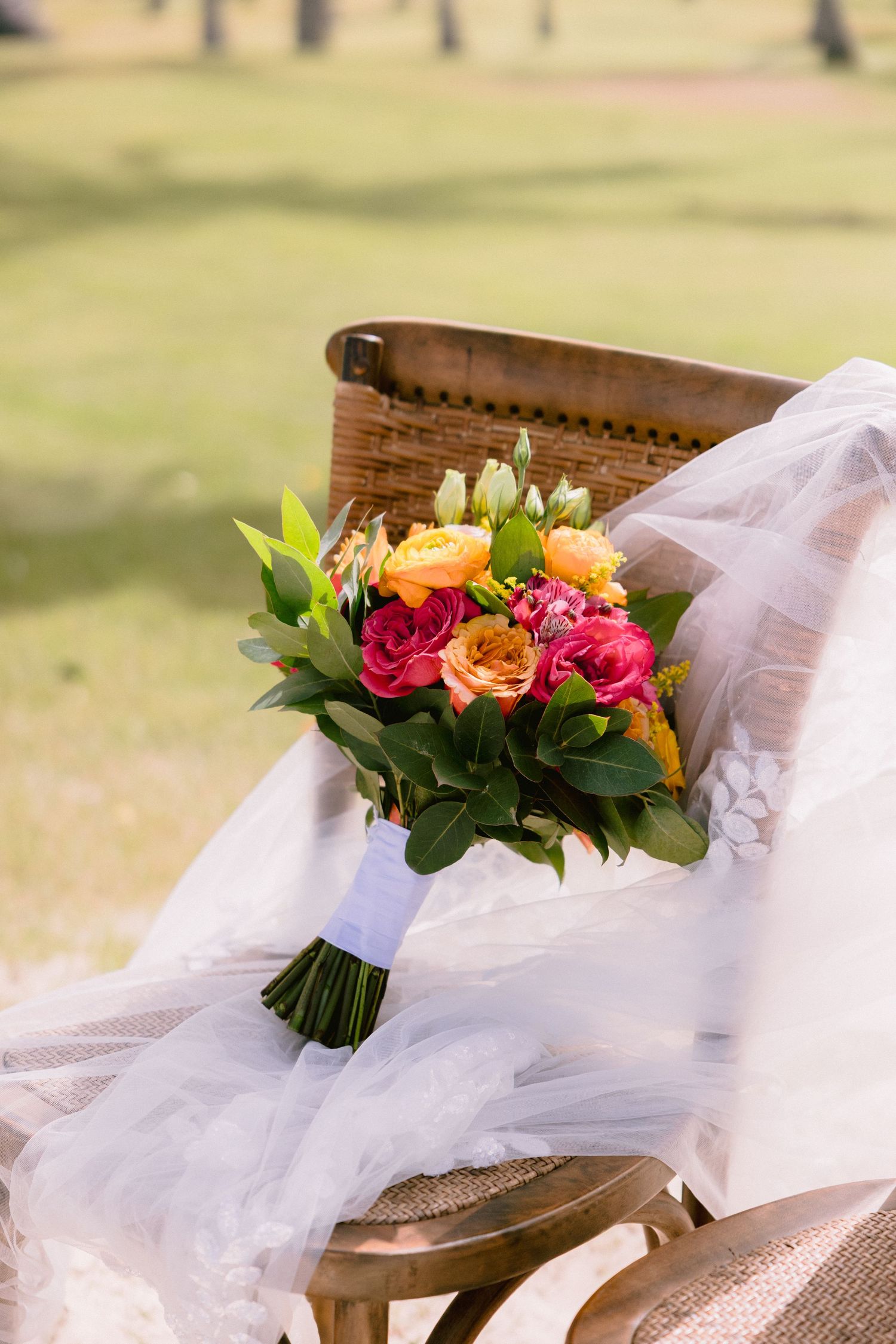 A vibrant bridal bouquet with pink and orange roses rests on white tulle fabric against a wooden chair outdoors.