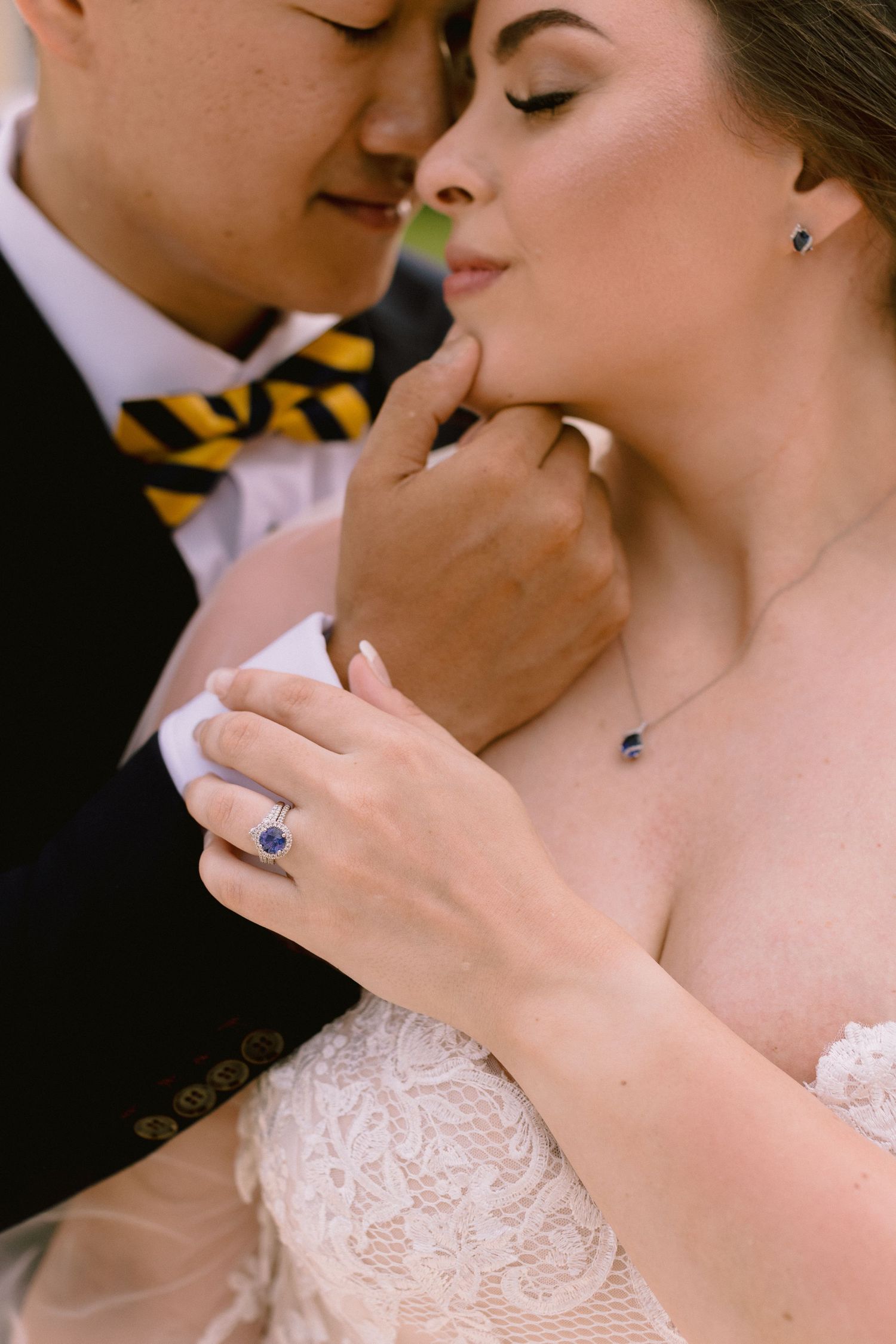 Bride and groom in close embrace, showcasing sapphire engagement ring, matching necklace and earrings, lace wedding dress.