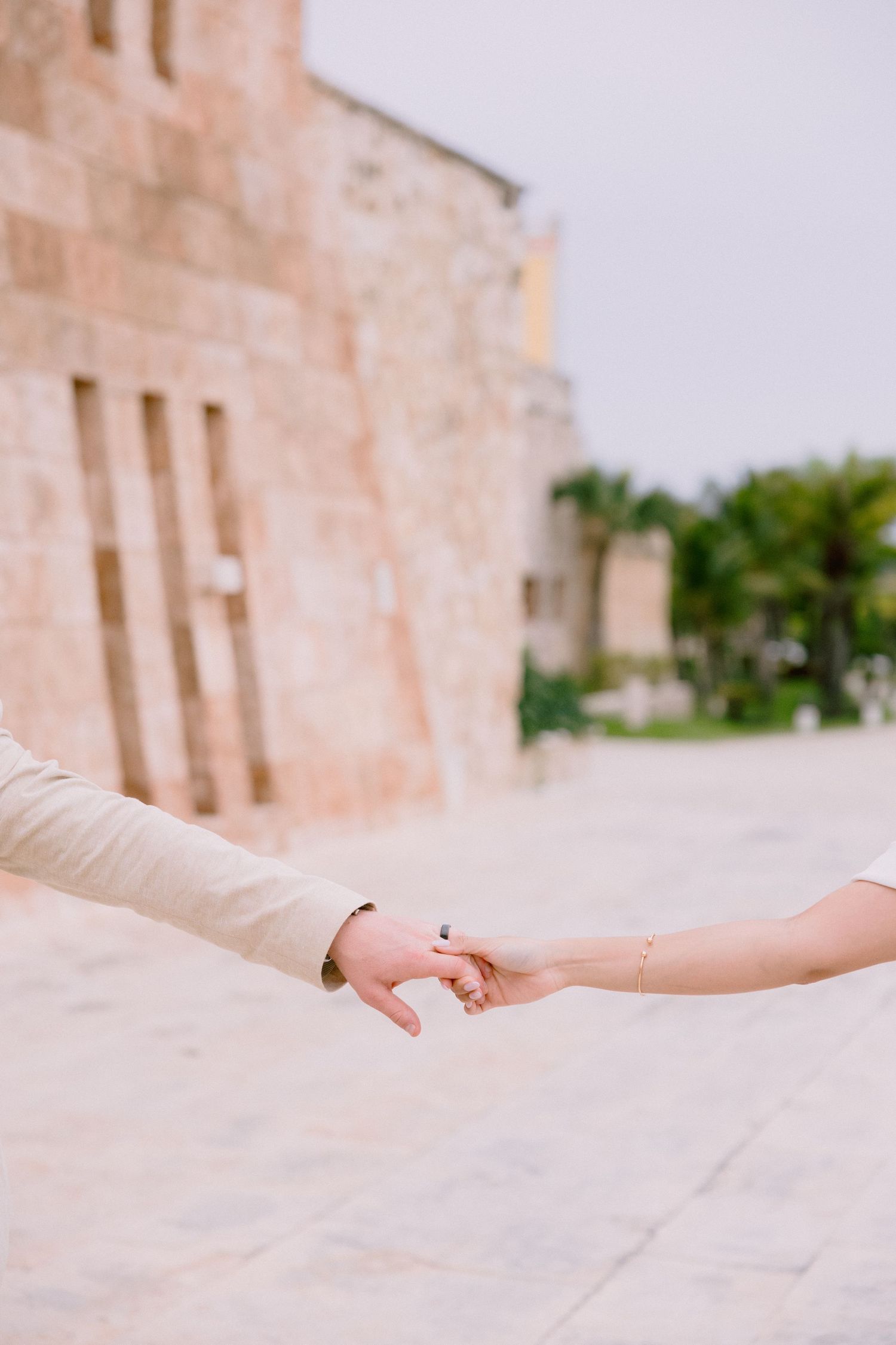 Couple holding hands tenderly near a historic stone building with palm trees in the background.