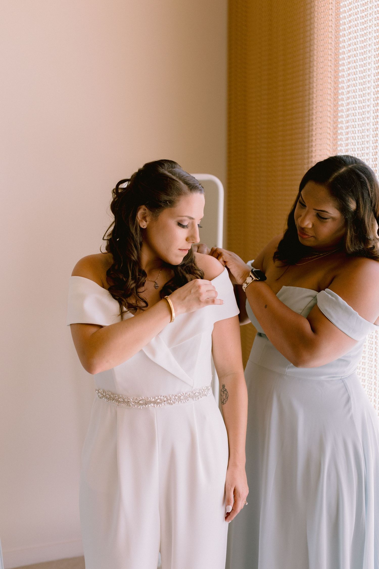 Bride in white off-shoulder jumpsuit being helped by bridesmaid in matching style near a window.