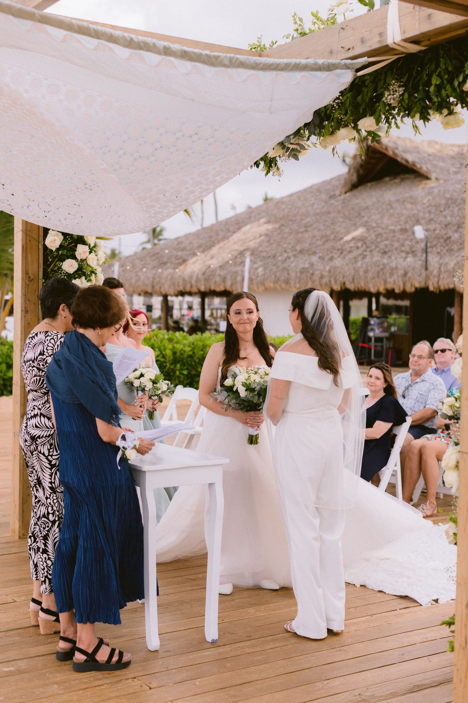 Two brides in white gowns exchange vows at an outdoor beach wedding ceremony under a rustic wooden pergola.