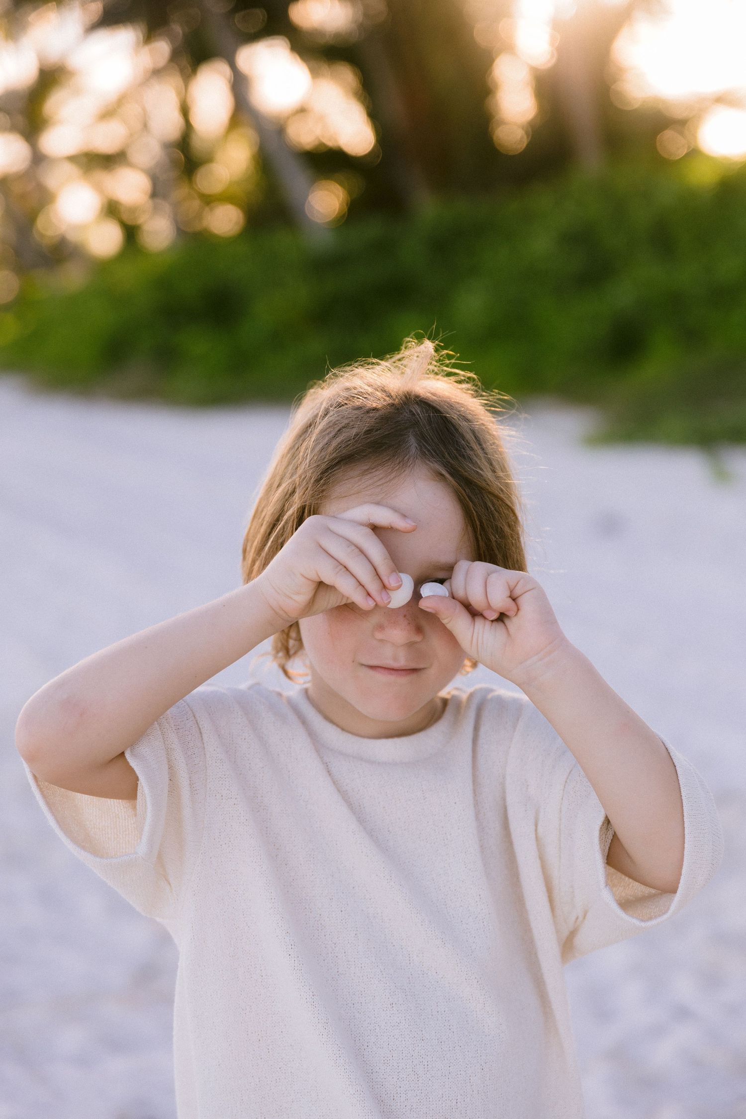 Young child rubbing eyes with both hands outdoors on sandy beach with green trees and golden sunset light.