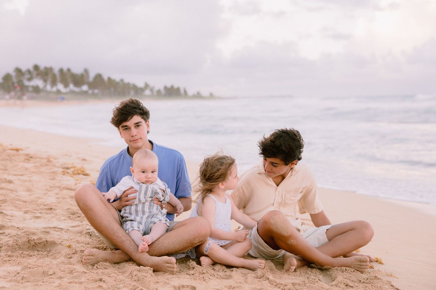 Two siblings sit on a sandy beach holding a baby and toddler with ocean waves in the background.