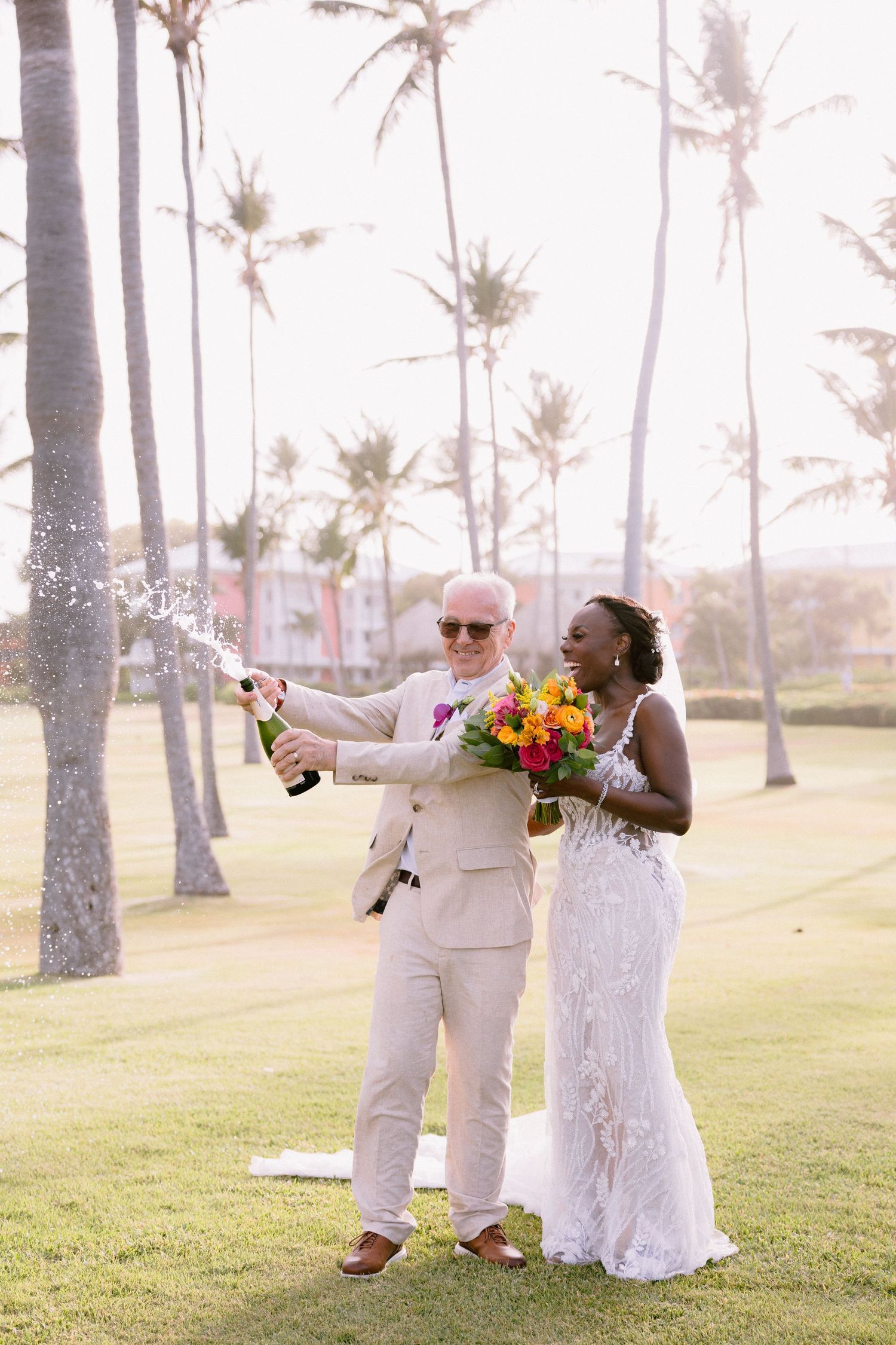 A joyful wedding couple celebrates with champagne on a palm tree-lined lawn during golden hour.