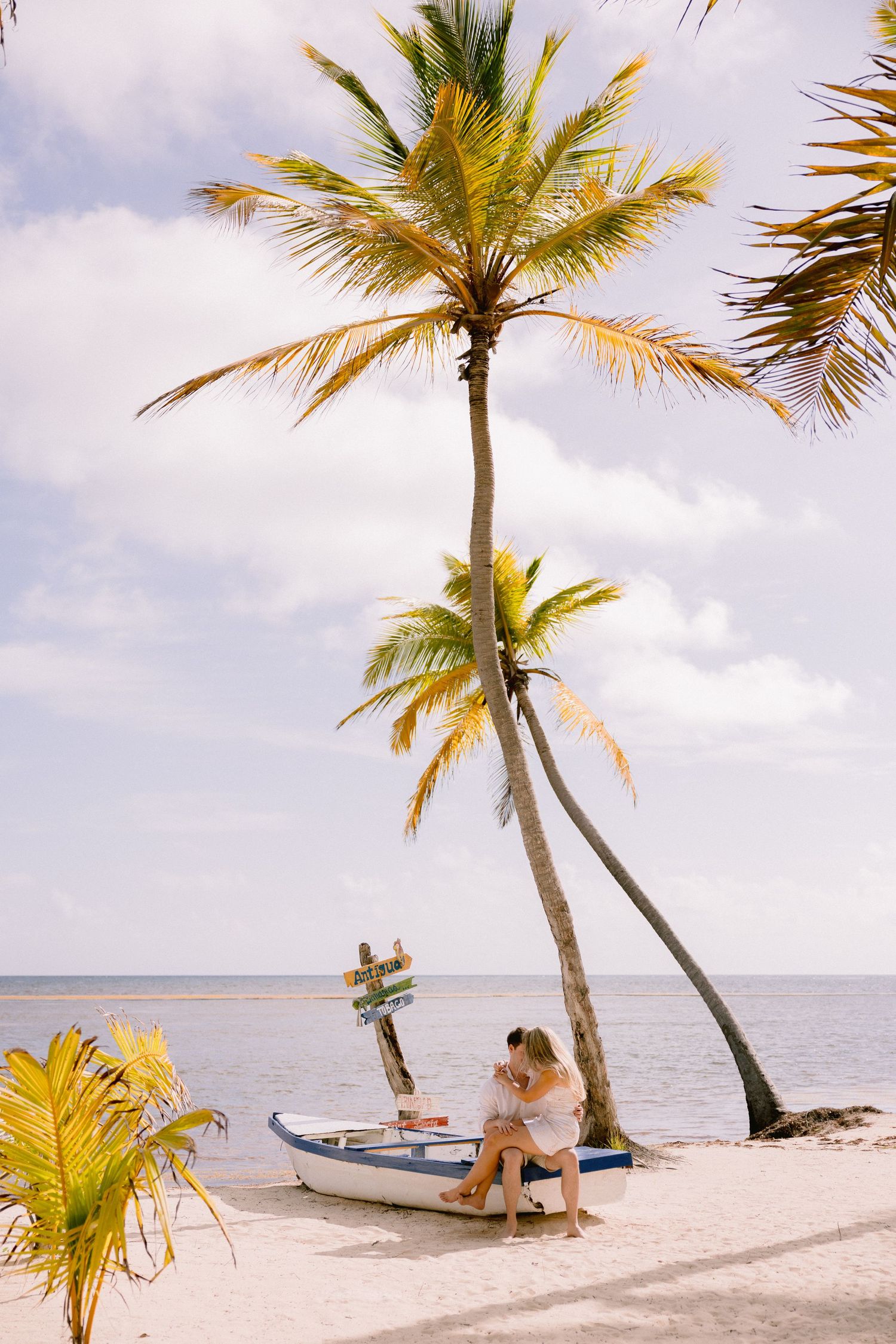 Romantic beach scene with palm trees swaying over a white sandy shore and small boat near the calm ocean at sunset.