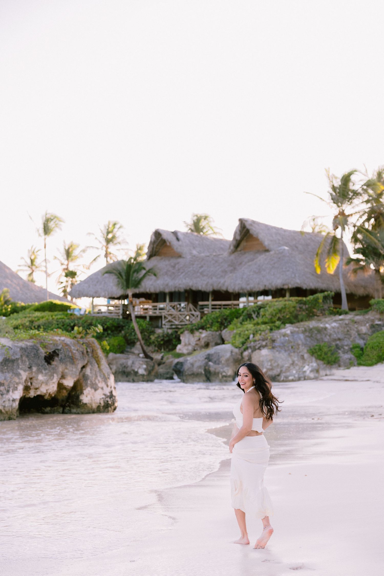 Woman in white dress walking barefoot on tropical beach with thatched huts and palm trees in background.