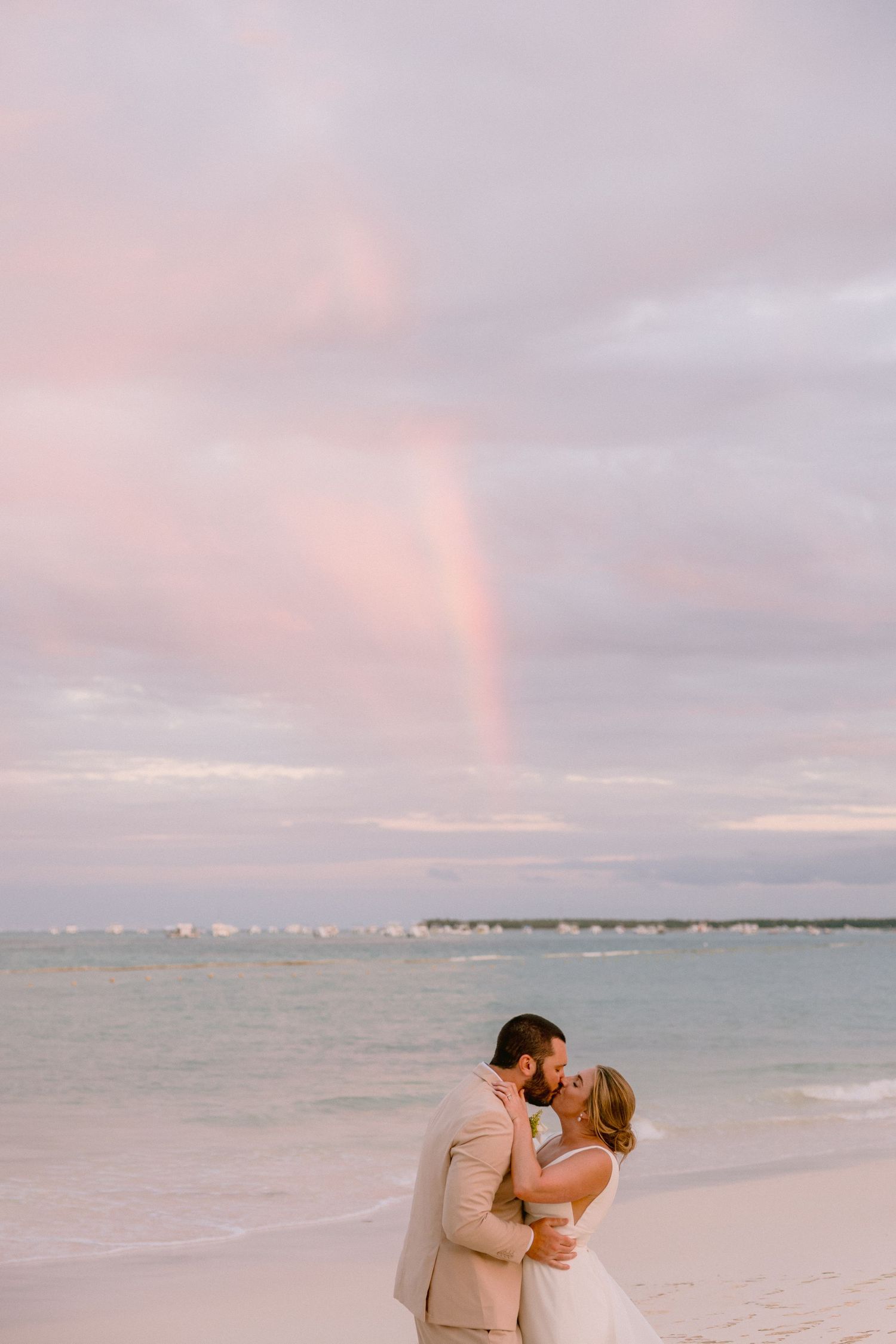 Bride and groom kissing on a beach at sunset with a rainbow in the pink sky over the ocean.