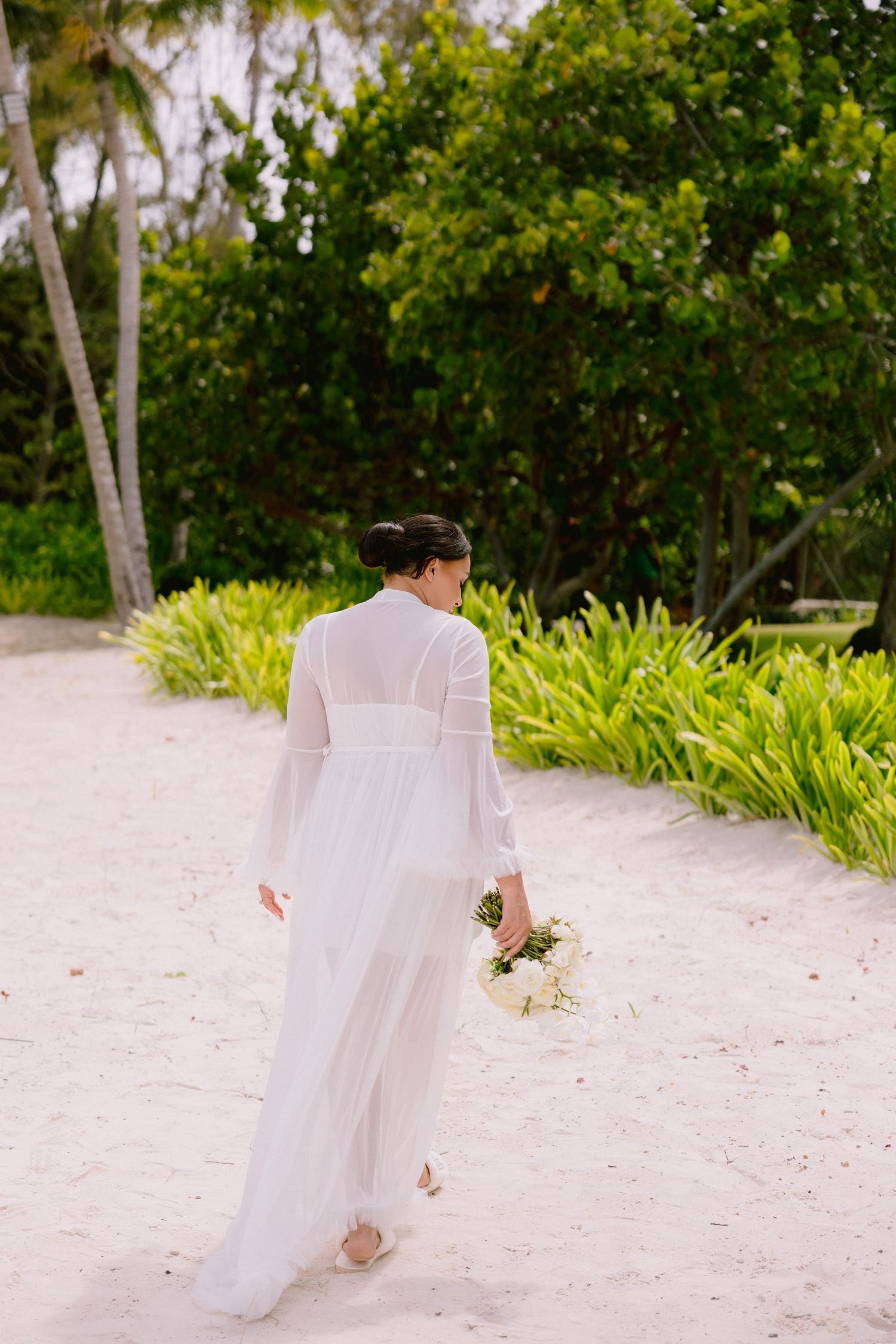 Bride in white long-sleeve gown holding bouquet walks barefoot on sandy beach path surrounded by lush tropical greenery.