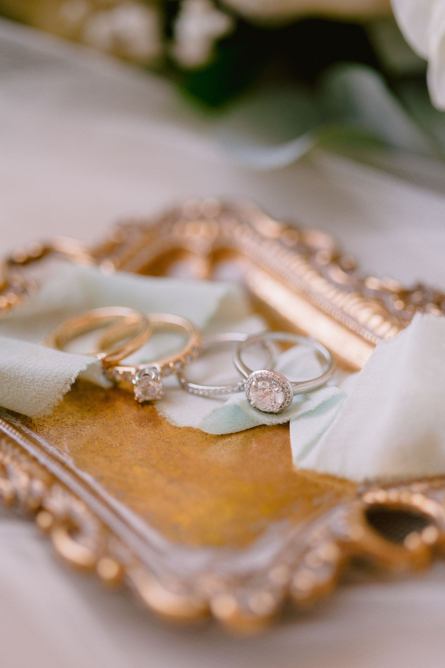 Elegant wedding rings displayed on a gold ornate tray with sage ribbon, featuring diamond halo and solitaire engagement rings.