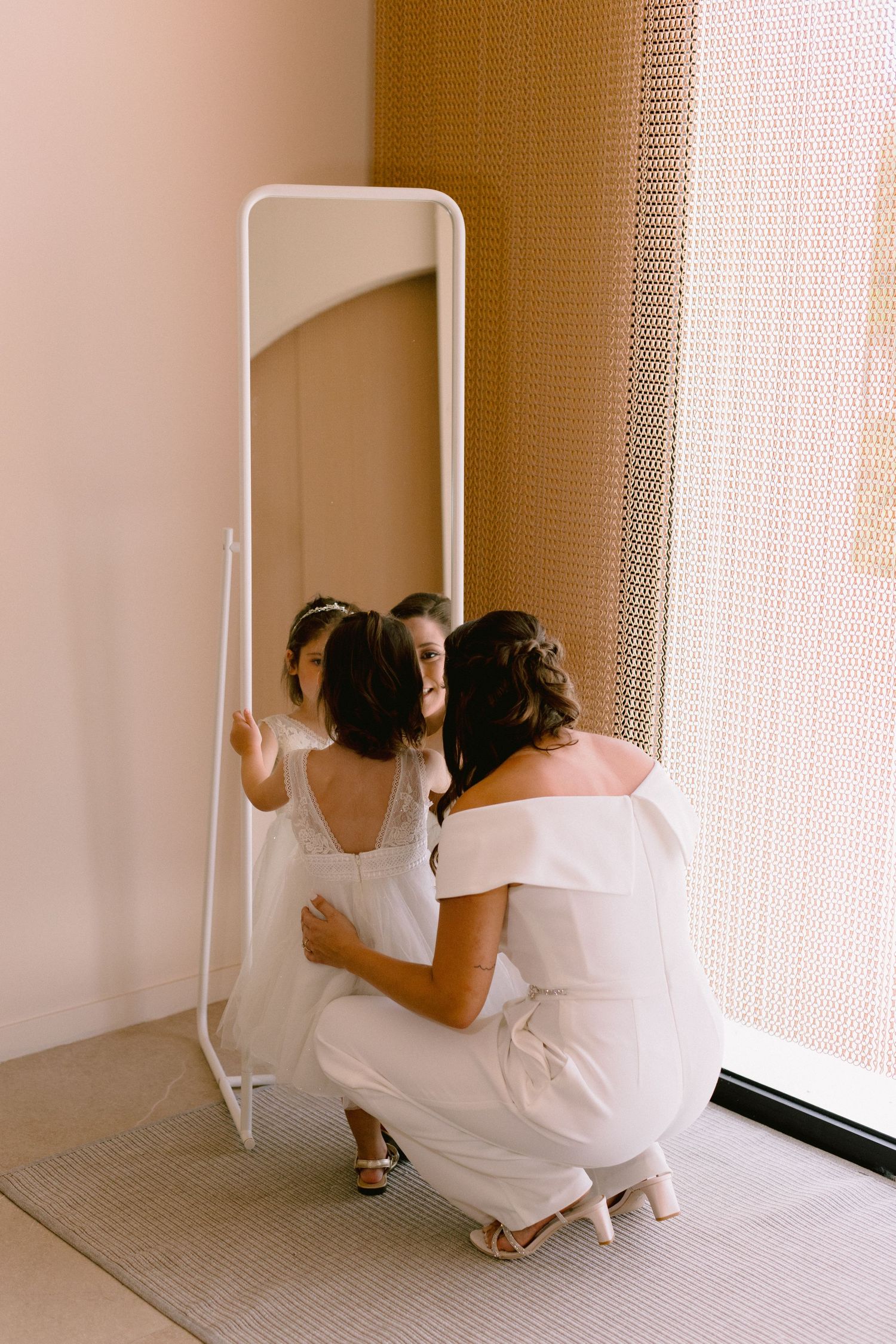 Bride and bridesmaid in white outfits crouch together before a full-length mirror, sharing a tender moment.