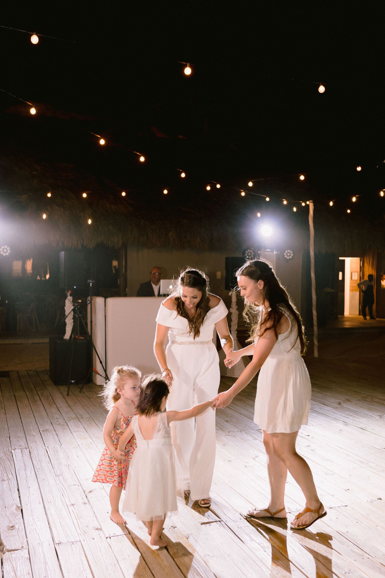 Bride dancing with bridesmaids and flower girls on wooden dance floor under string lights at outdoor wedding reception.