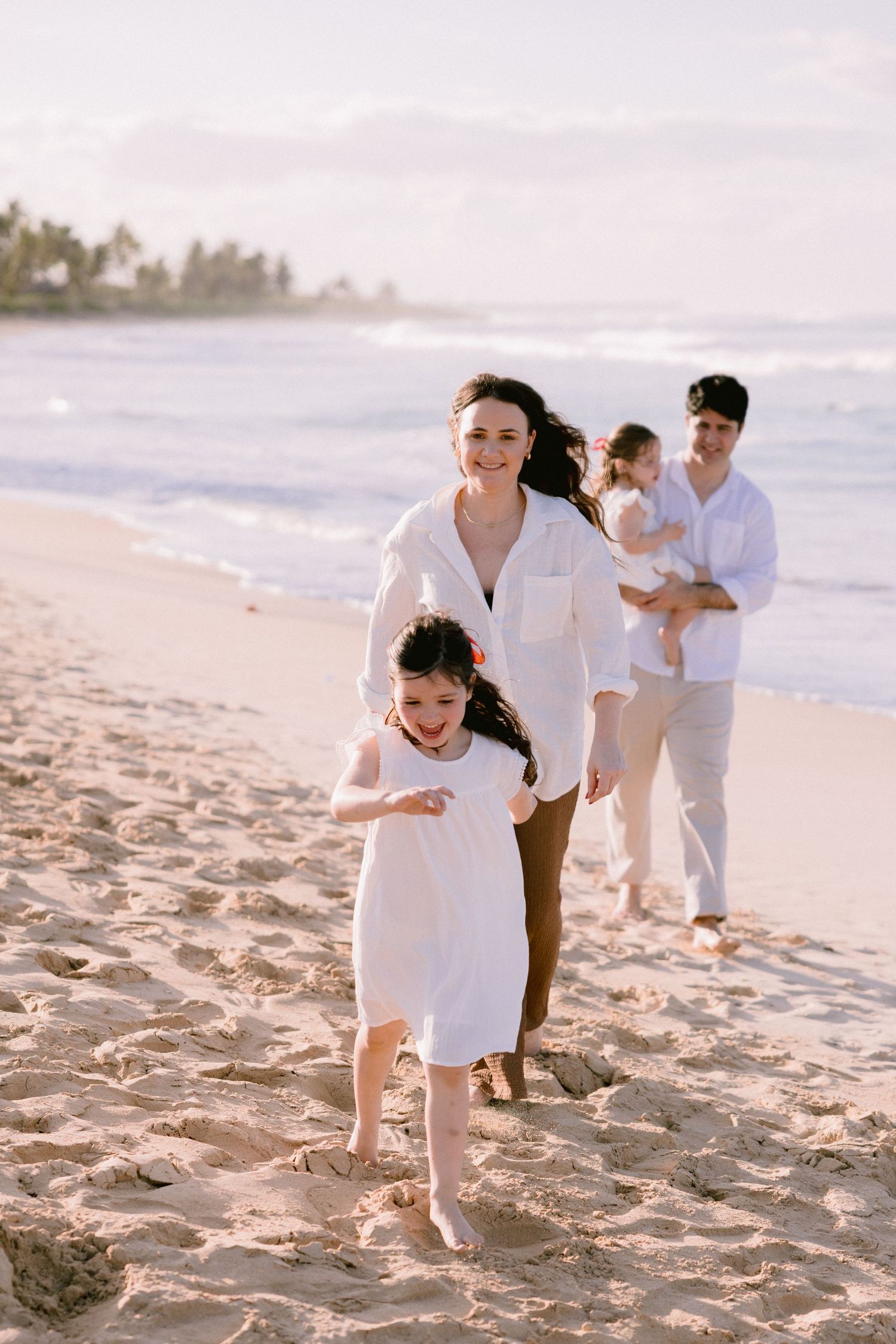 Family in white outfits playing and laughing together on a sandy beach during golden hour.