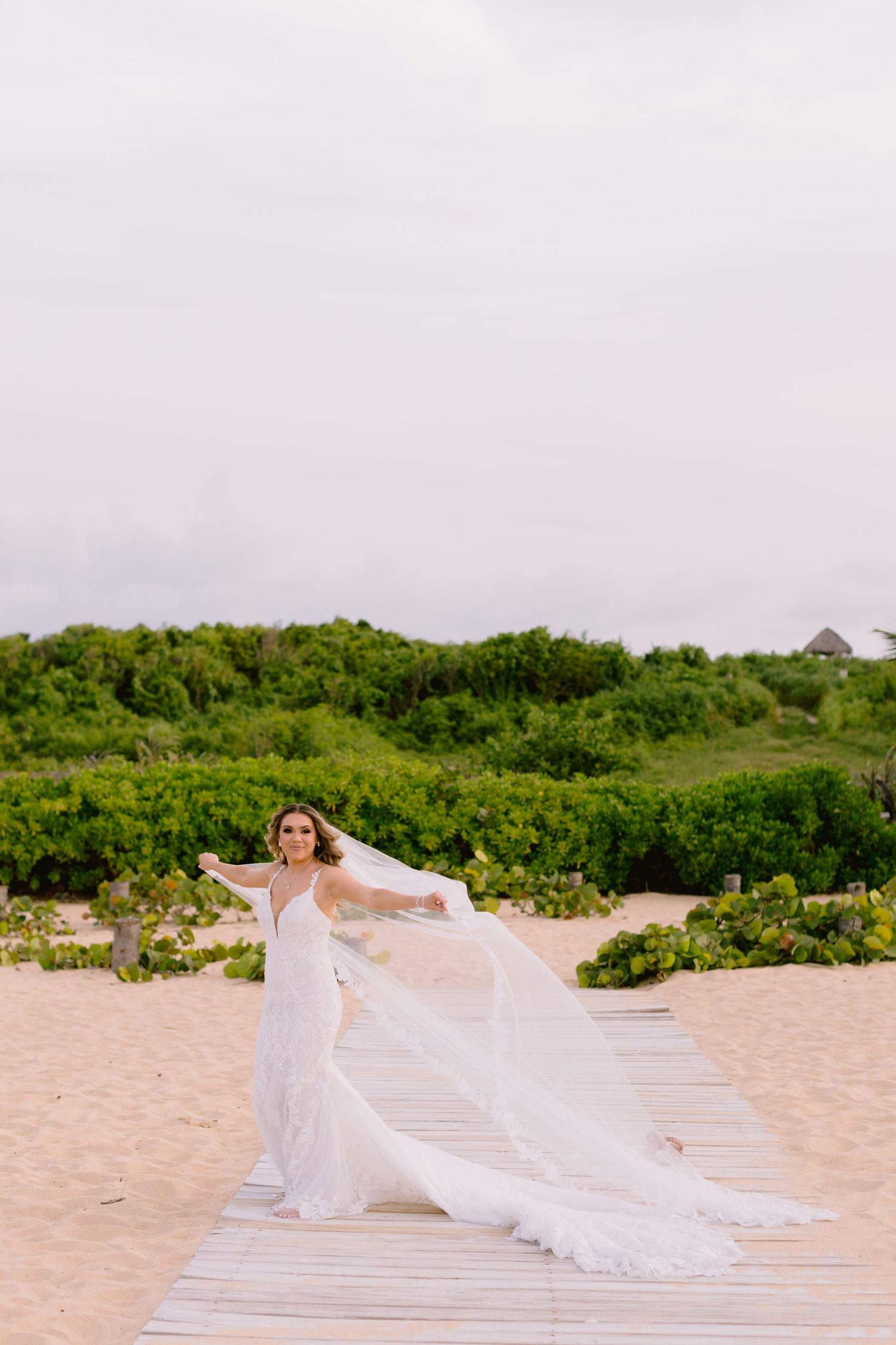 A flowing white wedding dress billows in the breeze as someone poses on a wooden boardwalk at a sandy beach.