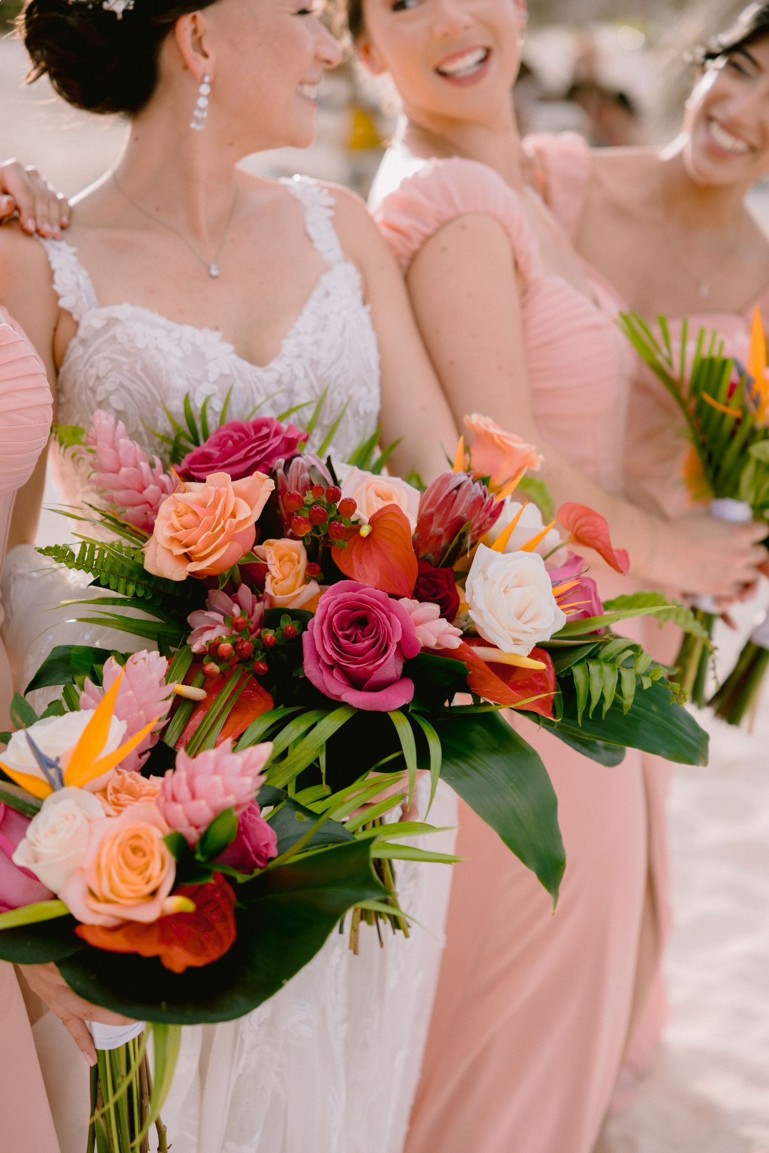 Vibrant pink and peach roses in wedding bouquet with tropical greenery complement bridesmaids in blush dresses.