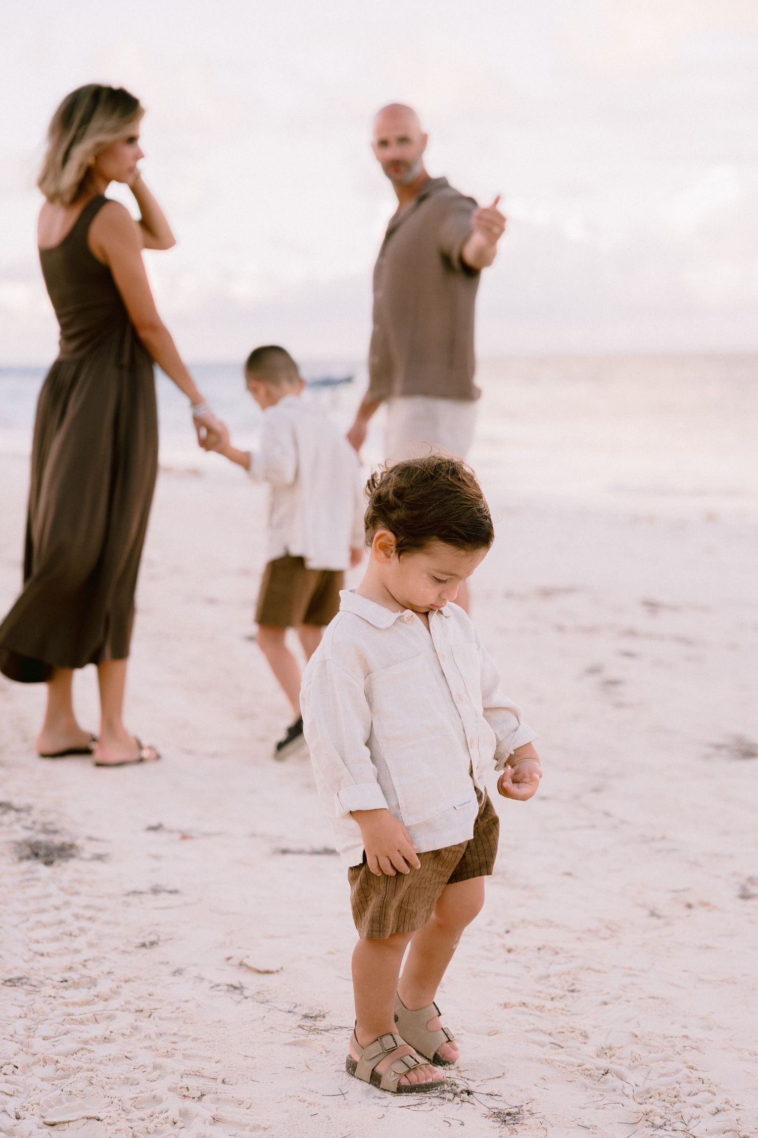 A child plays in the sand on a beach while people walk in the background during sunset.