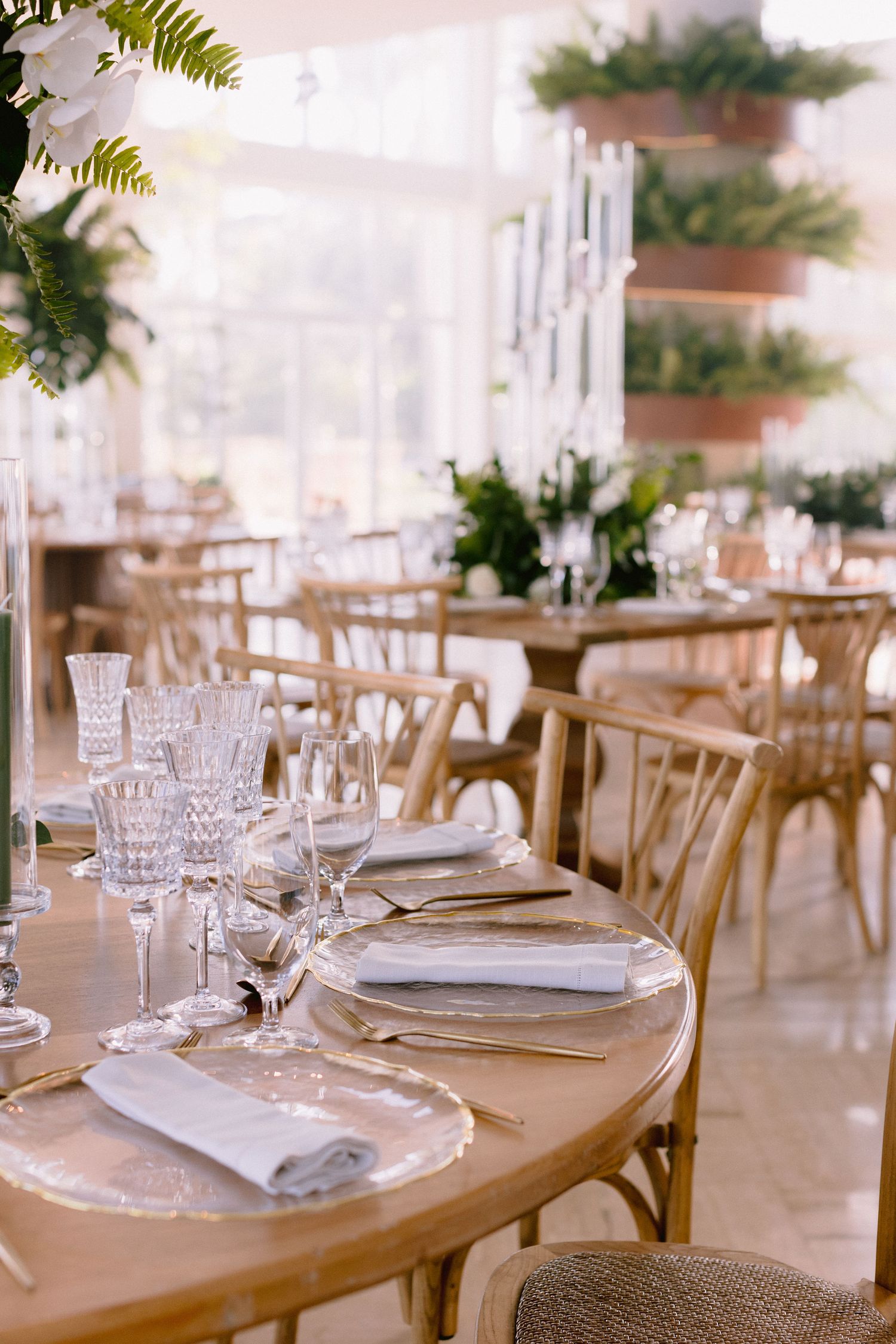 Elegant table settings with crystal glassware and white plates on wooden tables in a bright indoor reception space.