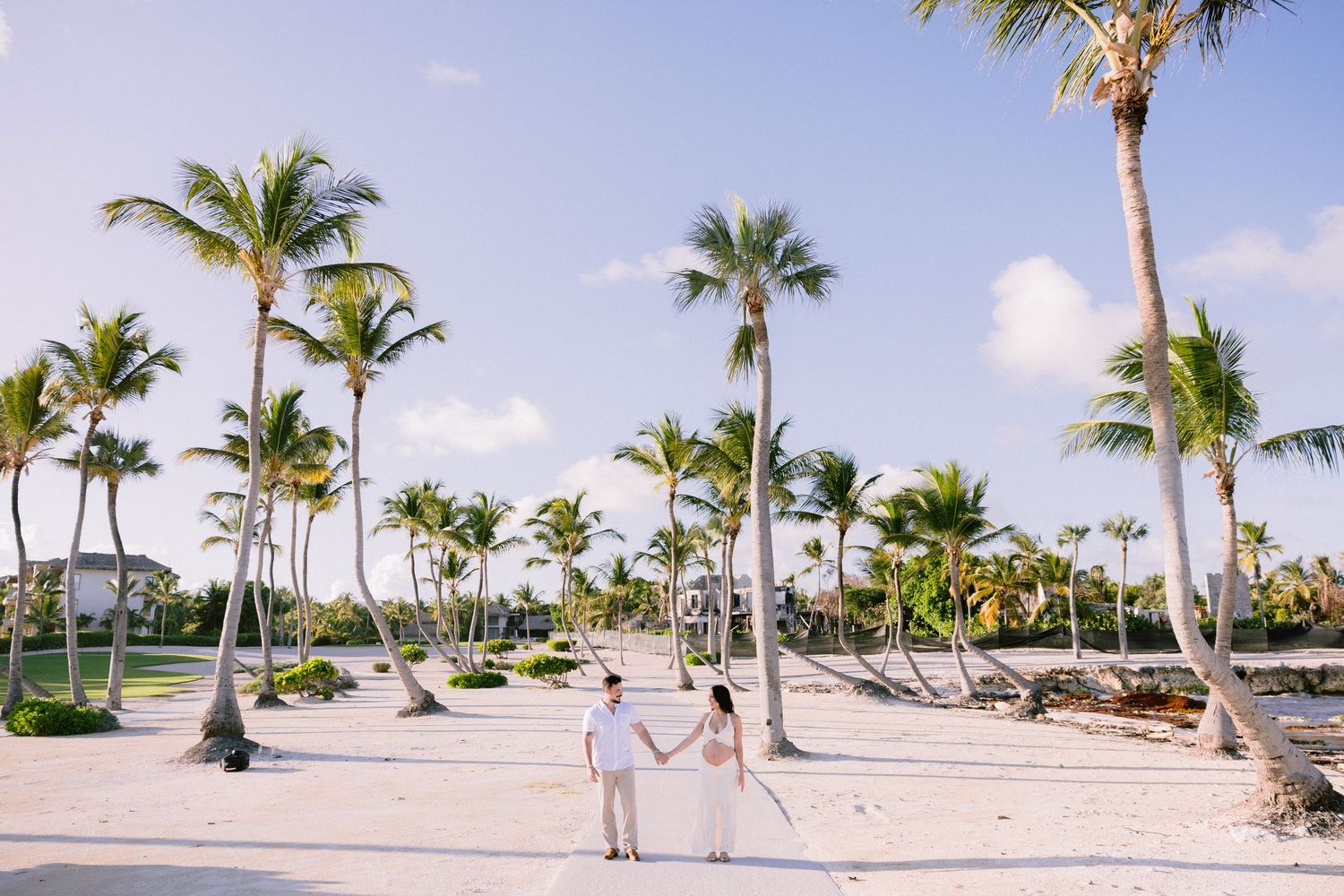 Couple holding hands on a white sand beach surrounded by tall palm trees at a tropical resort destination wedding.