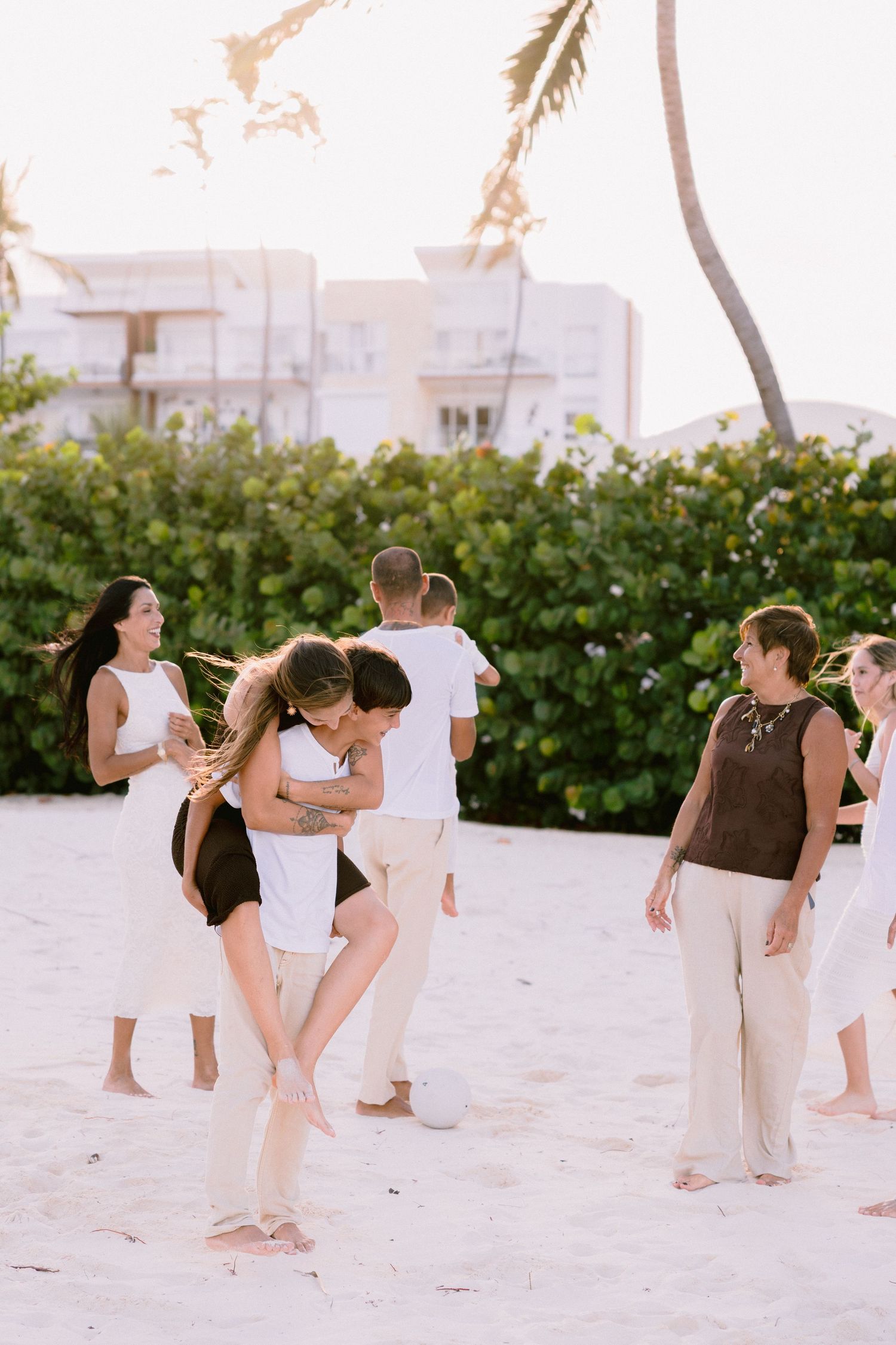 People in white clothing walk along a scenic beach with palm trees and hedges.