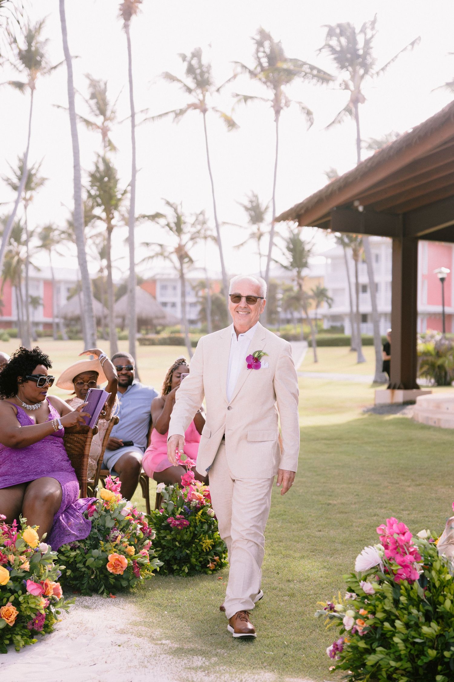 Wedding ceremony setup outdoors with colorful tropical flowers and palm trees lining a grassy pathway.