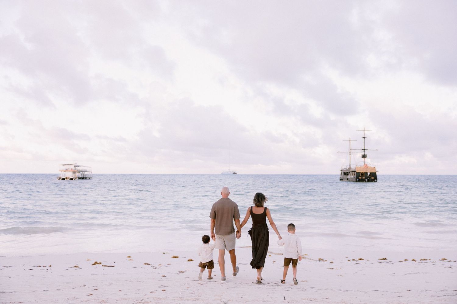 Family silhouettes walk along a white sandy beach at sunset with boats visible on the ocean horizon.