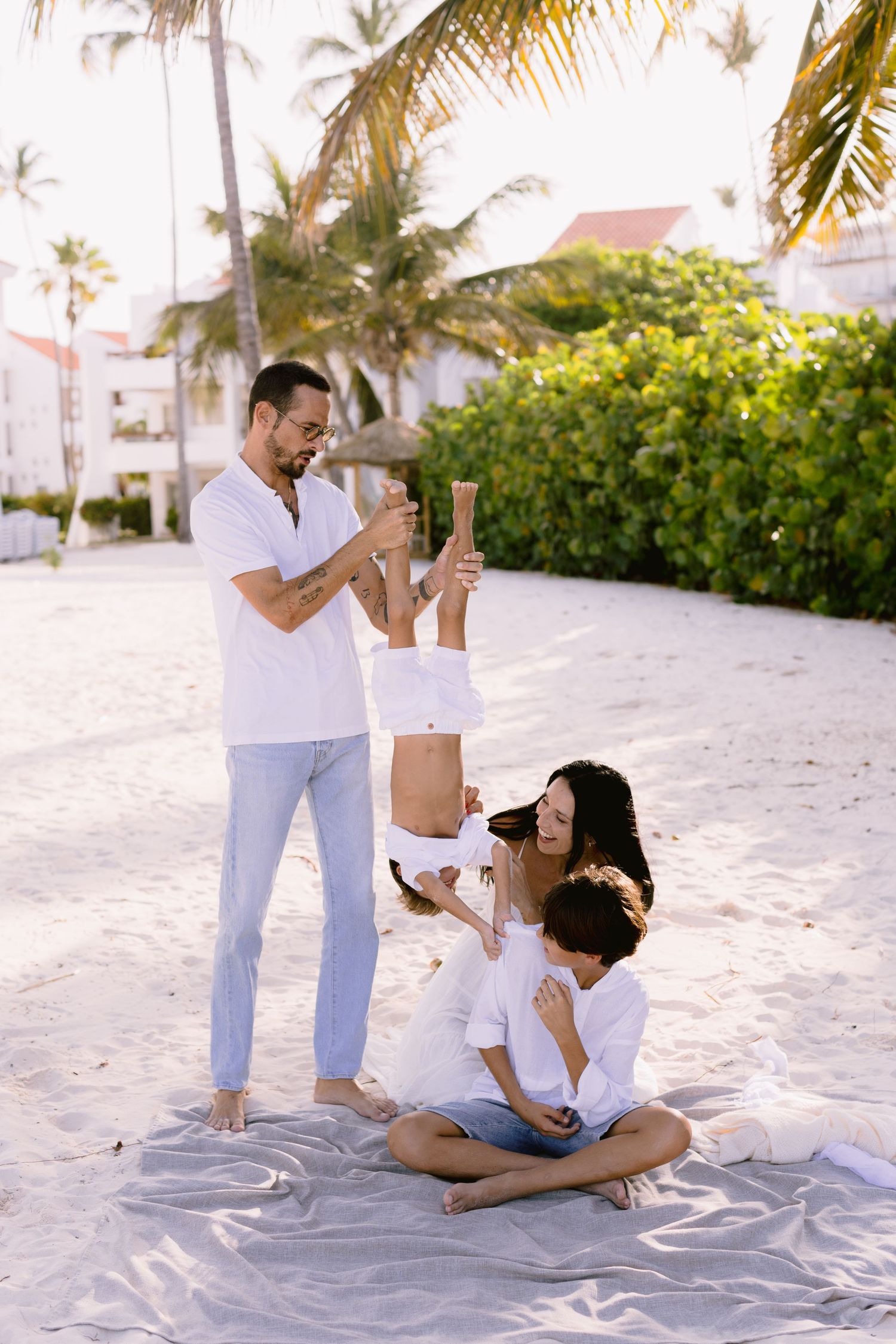 Family in white outfits enjoying a sunny beach day, with dad lifting child while mom sits on blanket with son near palm trees.