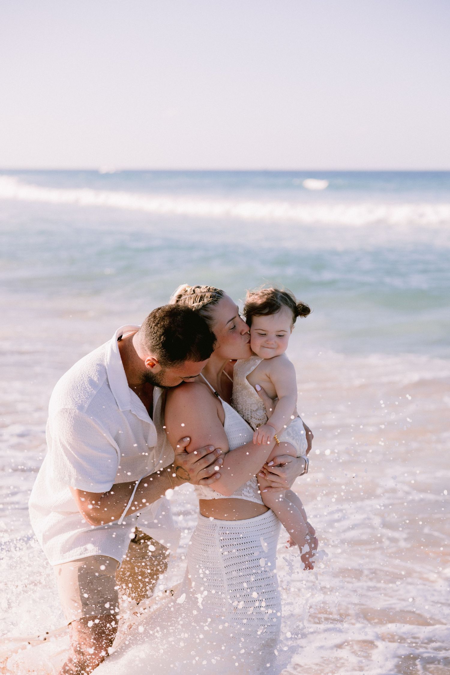 A happy family with a baby enjoys a sunny beach day, with waves splashing around them as parents kiss their child.