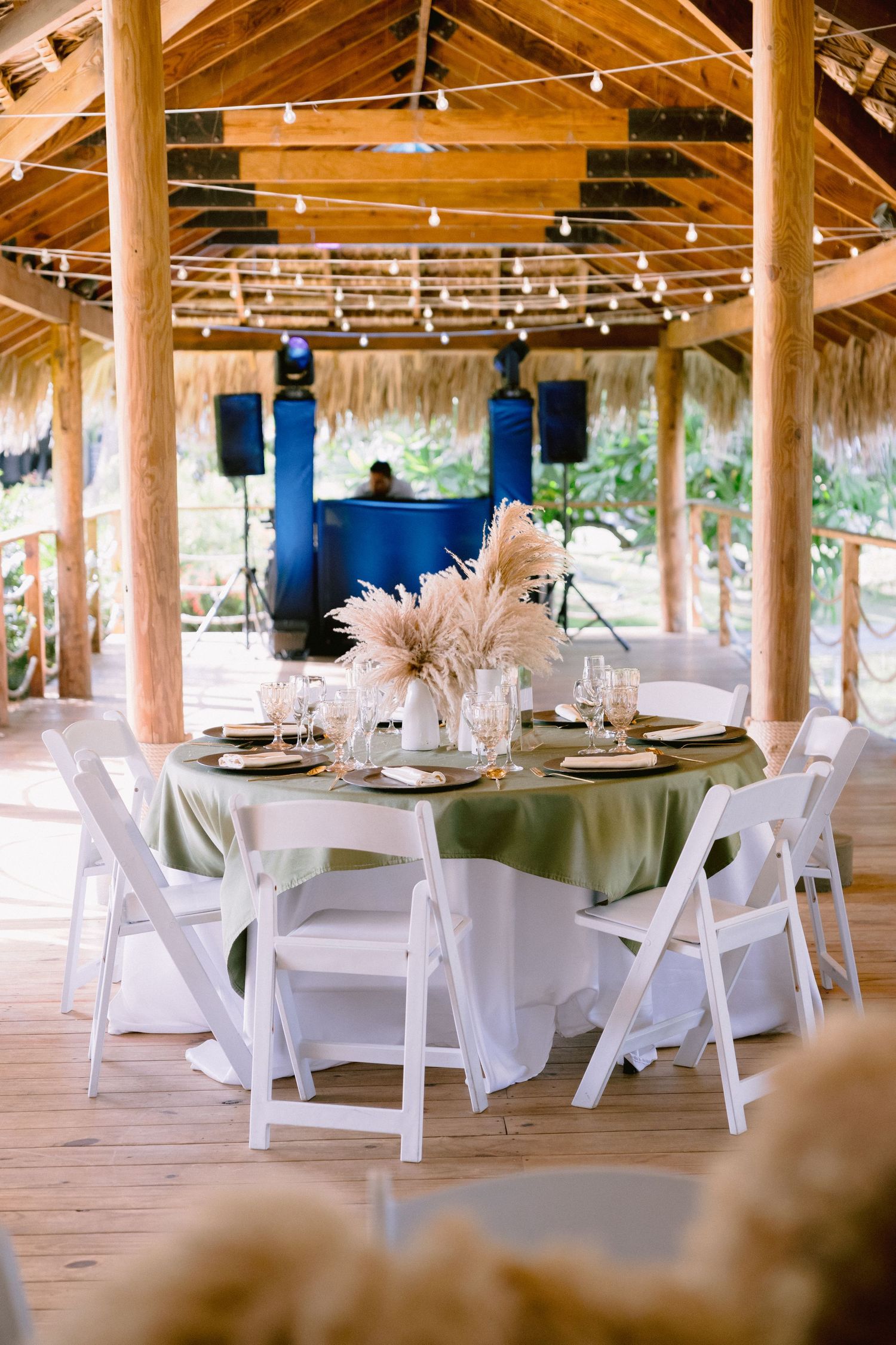 Elegant outdoor wedding reception under a thatched roof with white chairs, green linen tables, and pampas grass centerpieces.