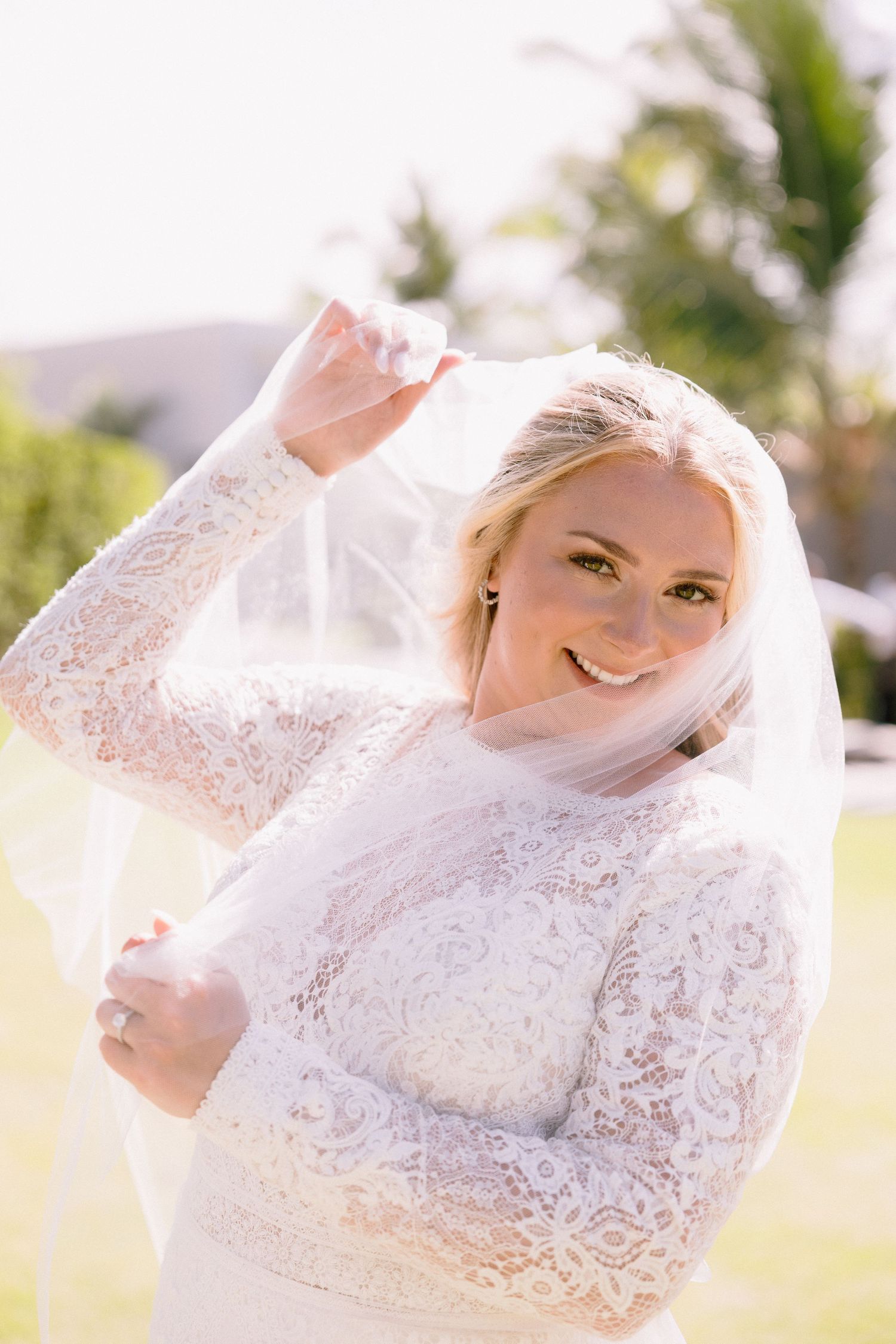 Smiling bride in white lace long-sleeve gown and veil poses outdoors on sunny day.