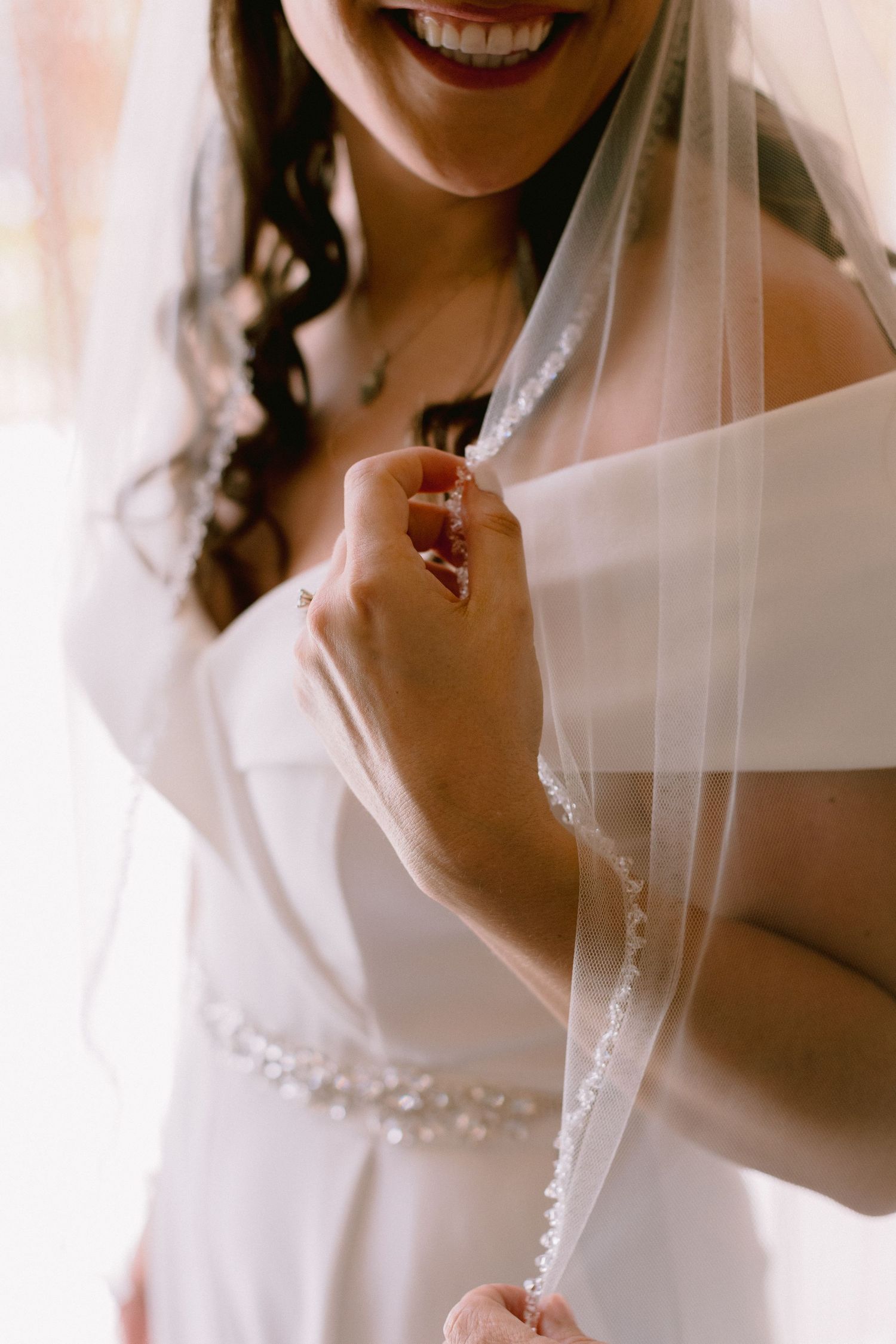 Smiling bride in white beaded gown holding her lace-trimmed veil, showcasing elegant wedding dress details.