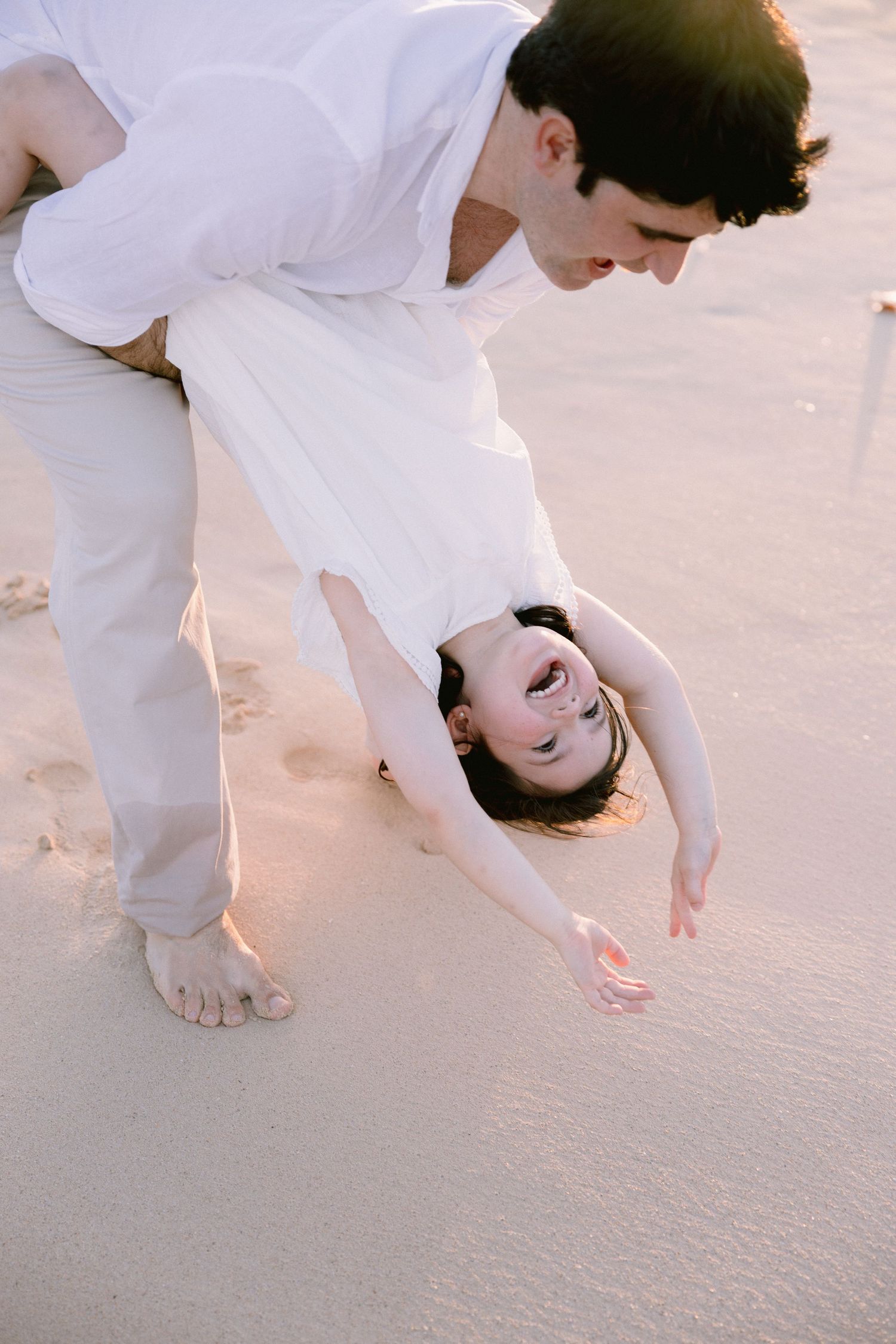 Person in white clothing bends down to touch sand on a beach at sunset, creating a peaceful coastal moment.