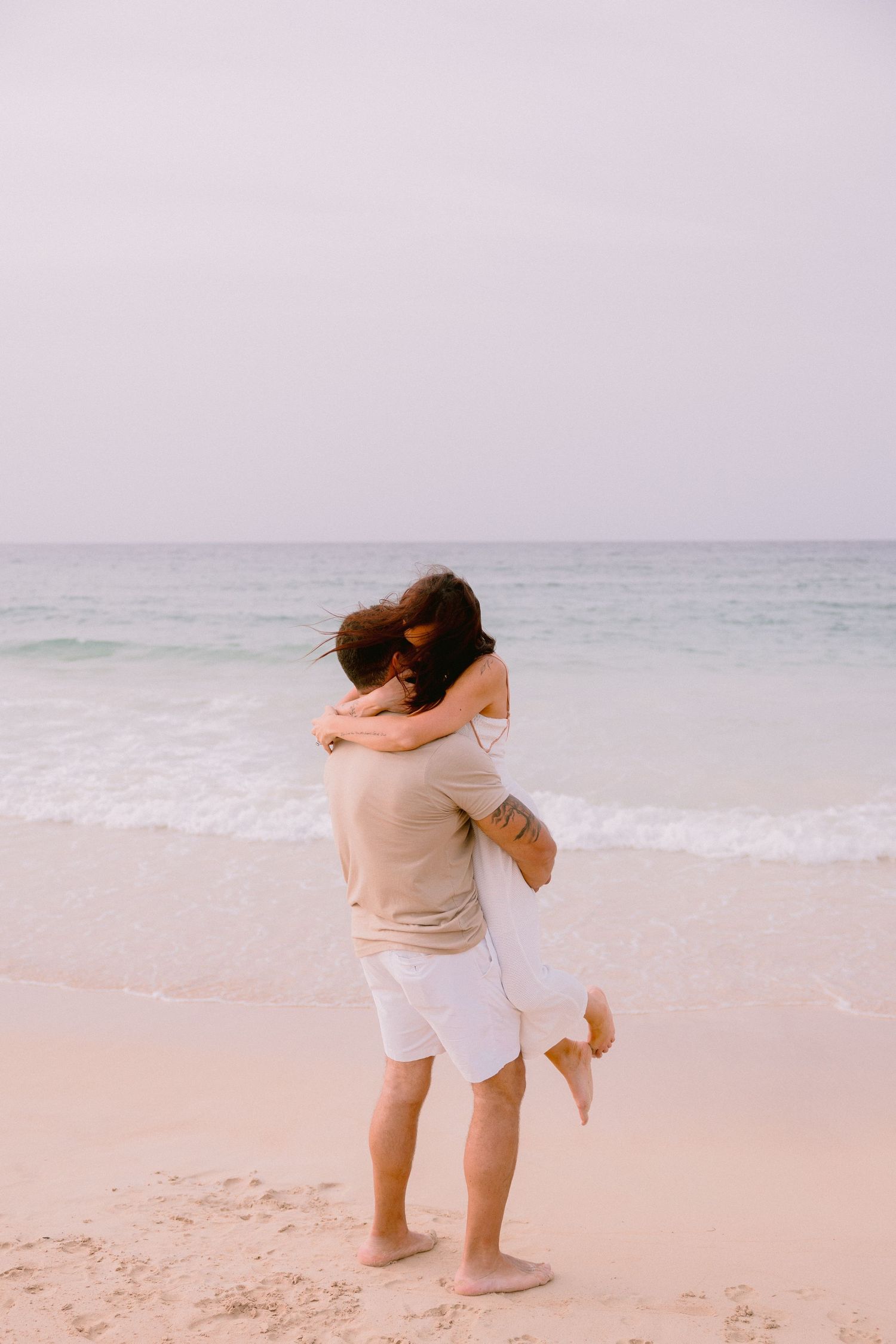 Couple embracing on a sandy beach with ocean waves in the background, wearing casual white and beige outfits.
